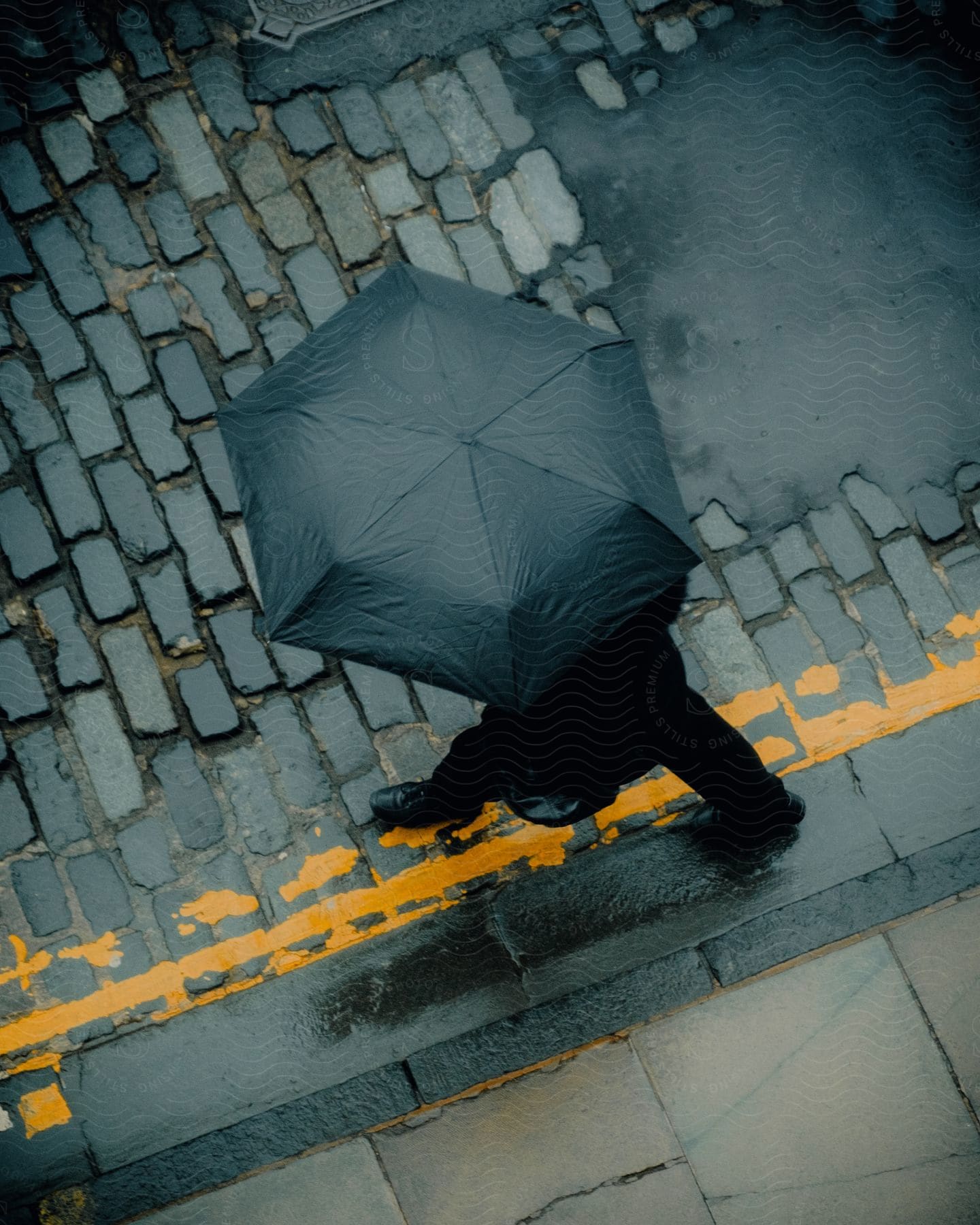 Top view of a person walking down a wet cobblestone street, wearing black formal clothes and an umbrella.