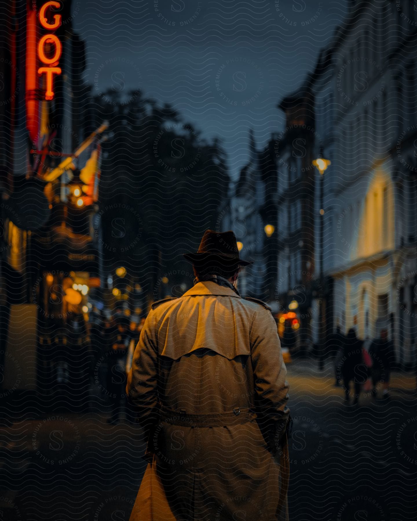 A man in a trench coat and hat walking down a city street at night, with illuminated signs in the background.