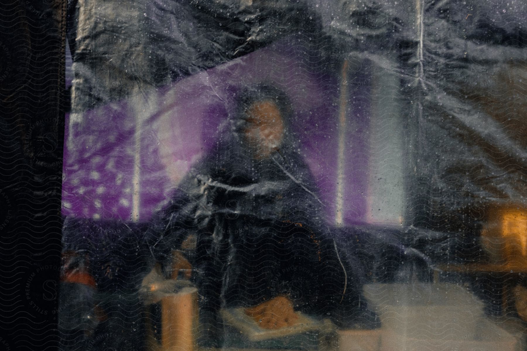 A woman, barely visible through the rain-dappled window, sits in front of a table wearing black clothing.