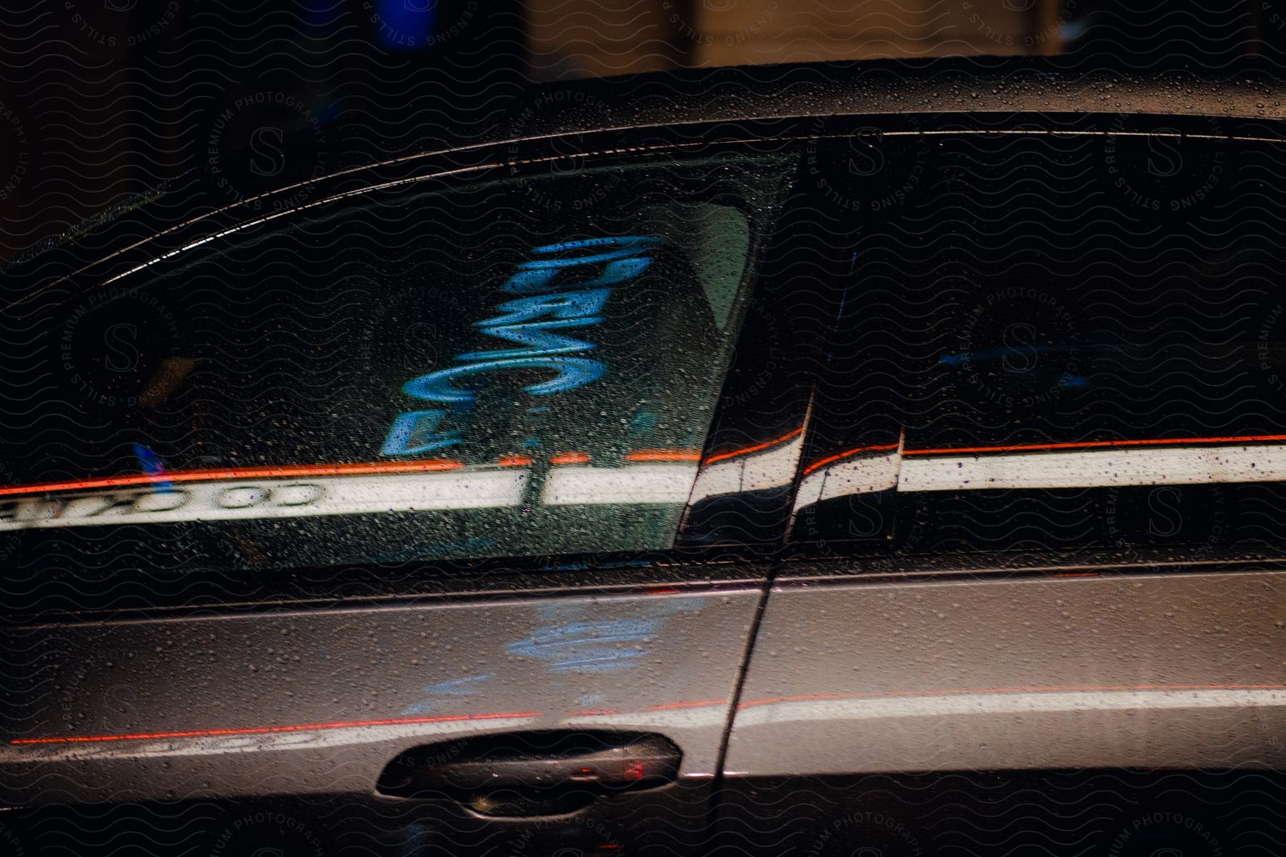 The body of a car, seen from the driver's side door and window, reflects light at night, with rain droplets covering the vehicle.