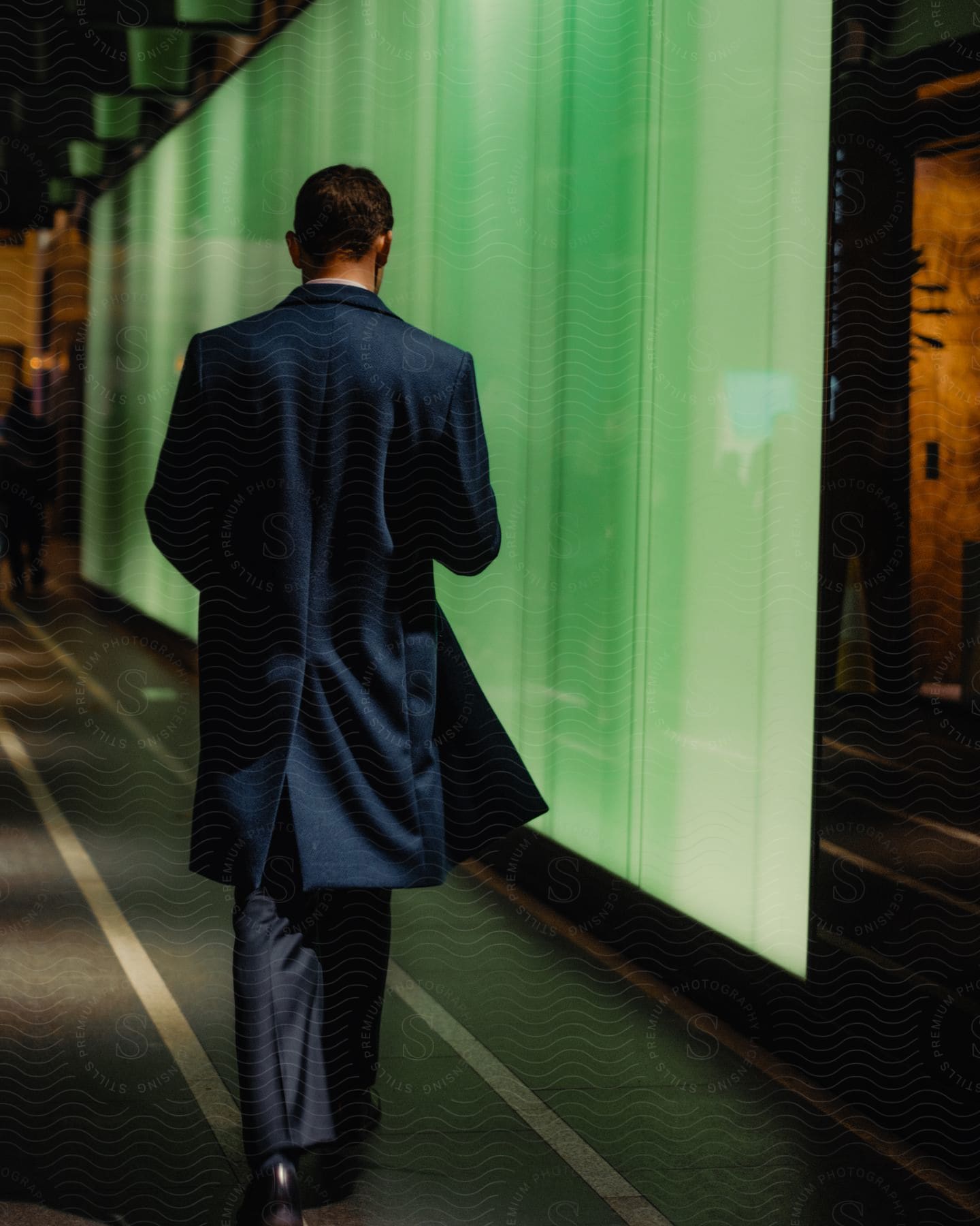 Man in a navy coat walking past a glowing green wall in a modern, dimly lit corridor.