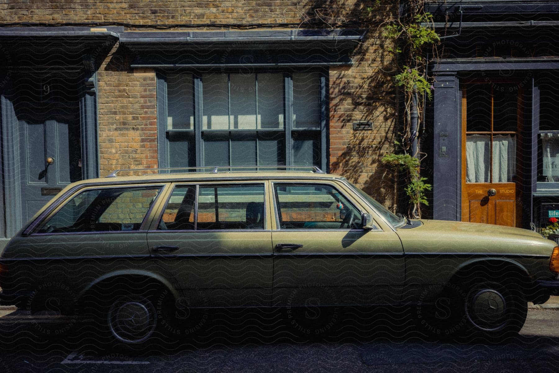 An olive-colored vintage station wagon is parked in front of a brick building with two doors and one window in the suburbs.