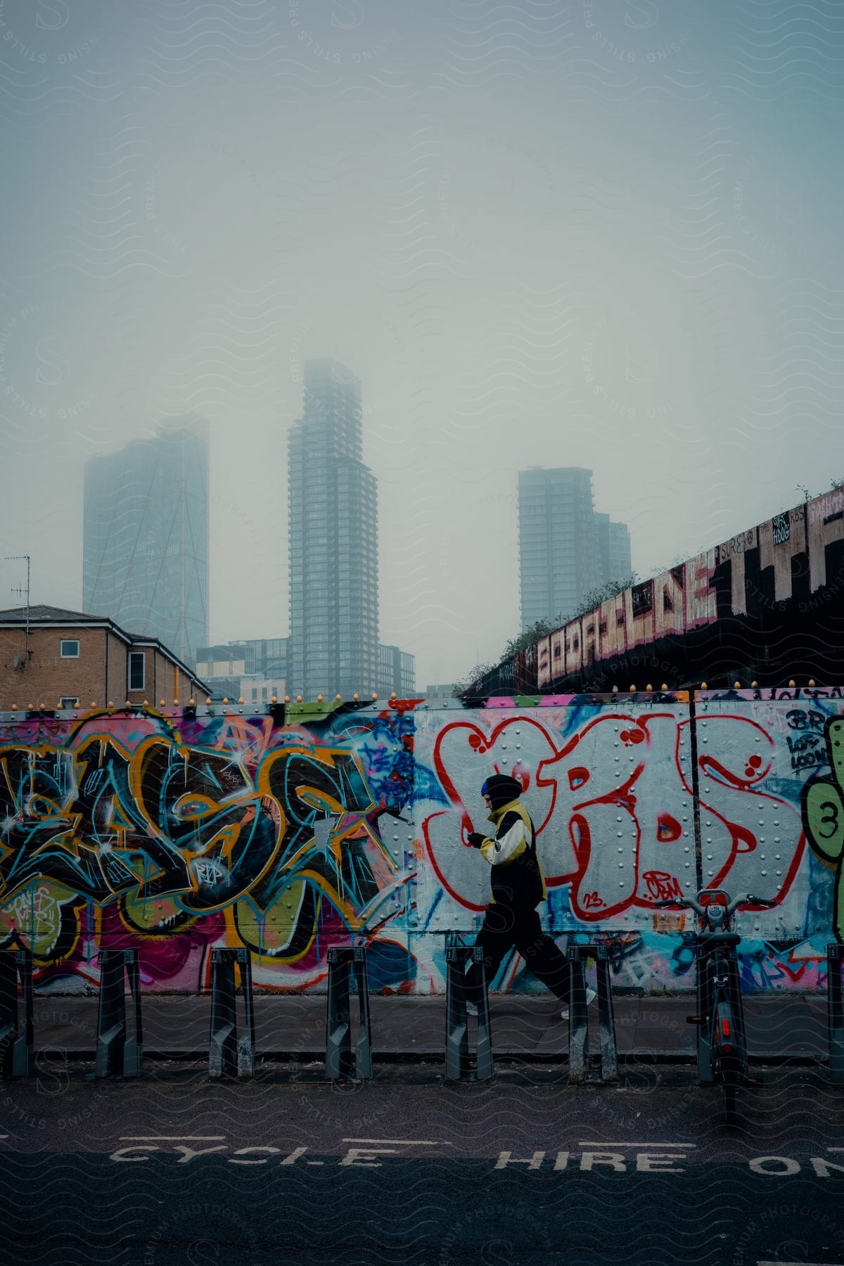 A person walks past a wall of graffiti in a foggy, urban area.