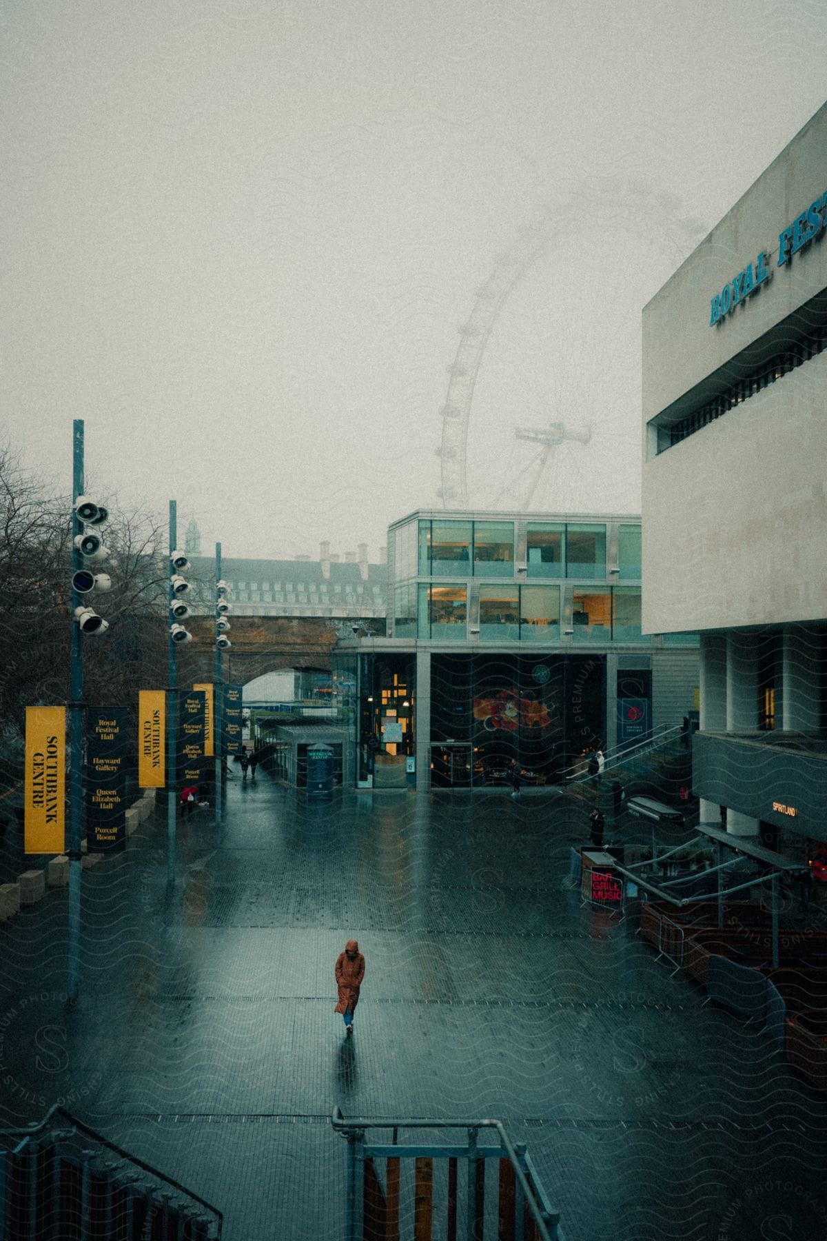 People walk on a wet paved area near a building, with a Ferris wheel visible in the background through the fog. The environment has a cold and gloomy tone.