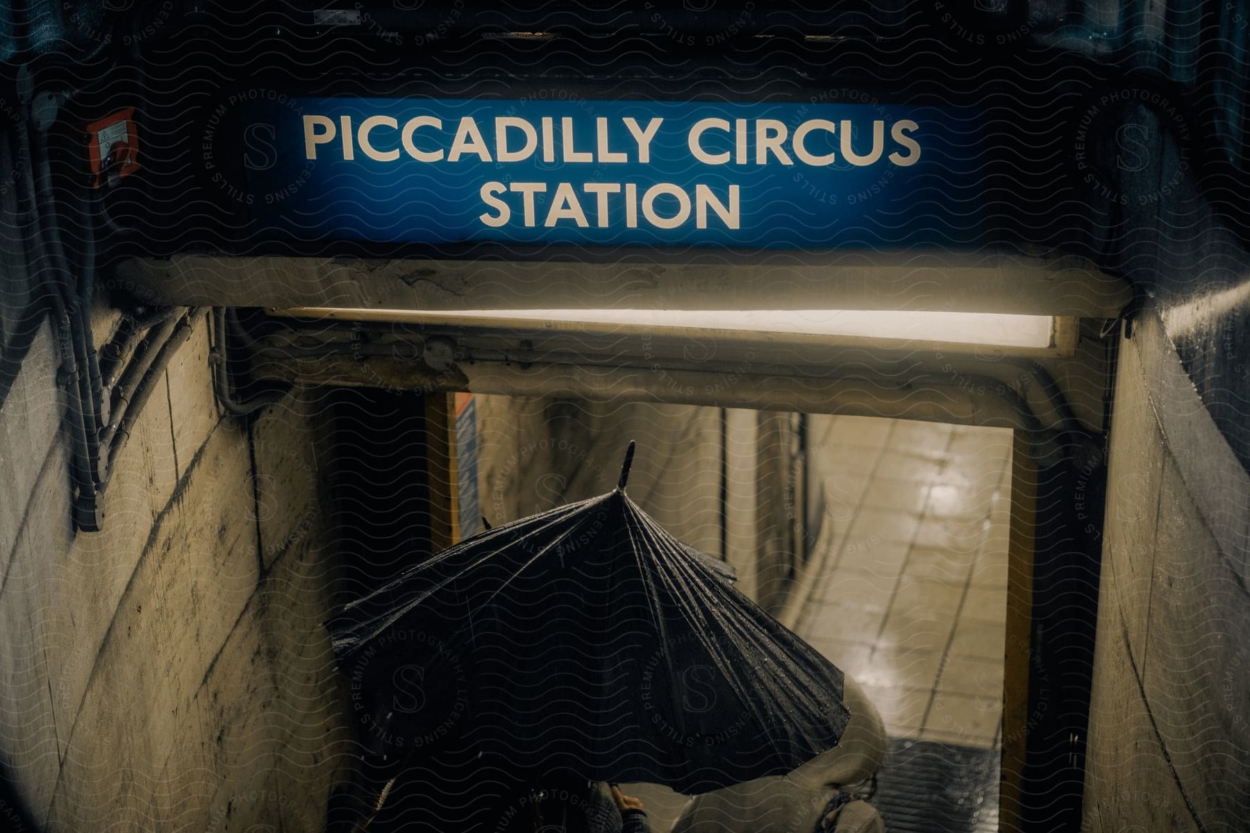 A person entering a subway station using a black umbrella and there is a blue sign at the entrance