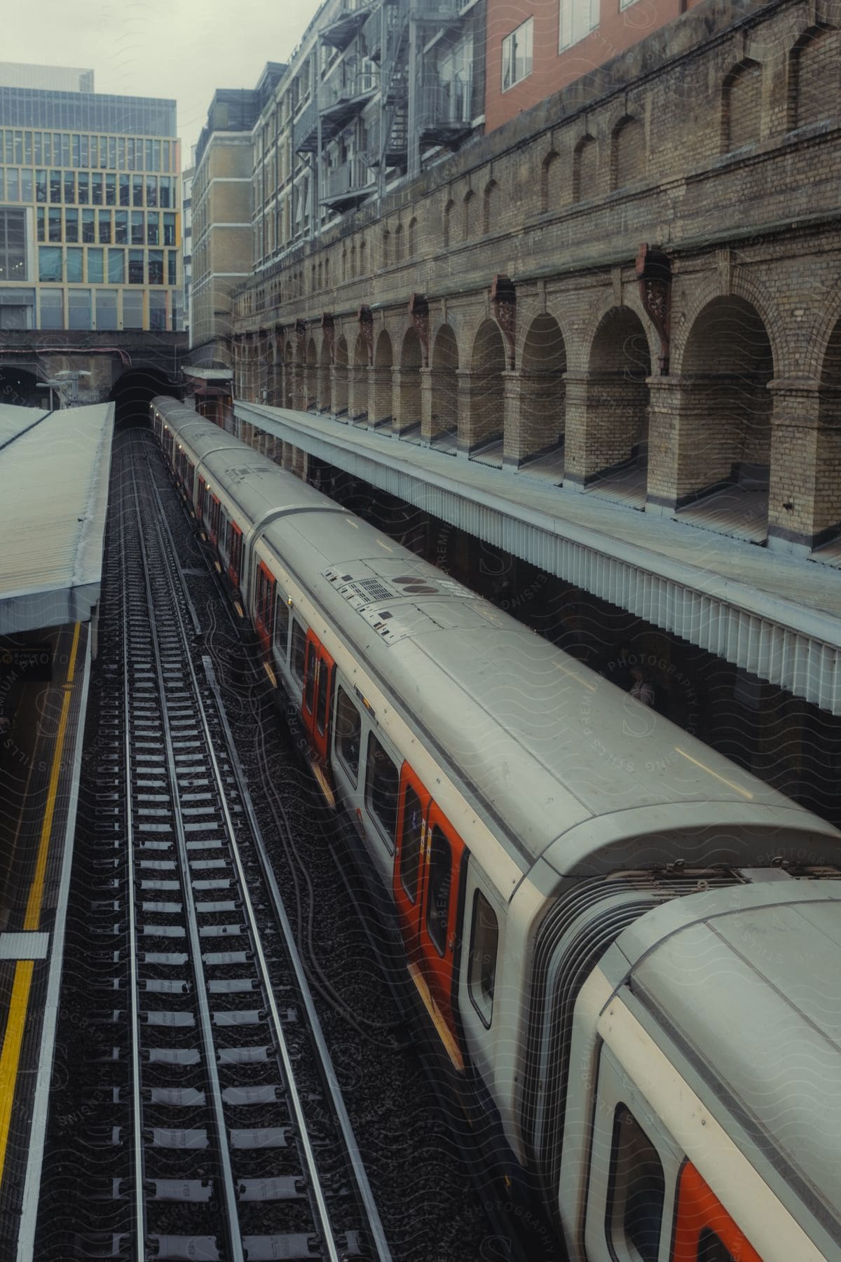 Train at platform in urban railway station with arched brick structure and adjacent buildings.