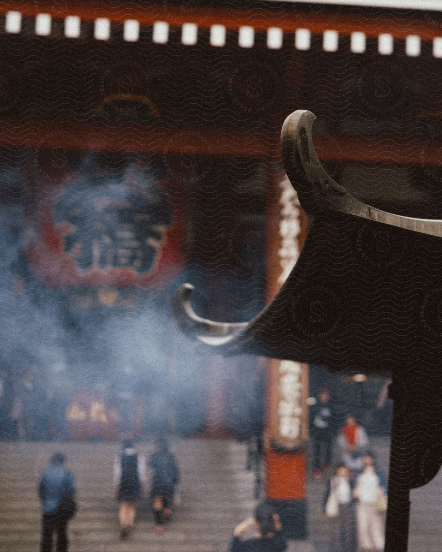 A large incense burner emits smoke in front of a traditional building, while people circulate around it. The atmosphere is serene and spiritual, with the incense and the architectural style of the building contributing to this feeling.