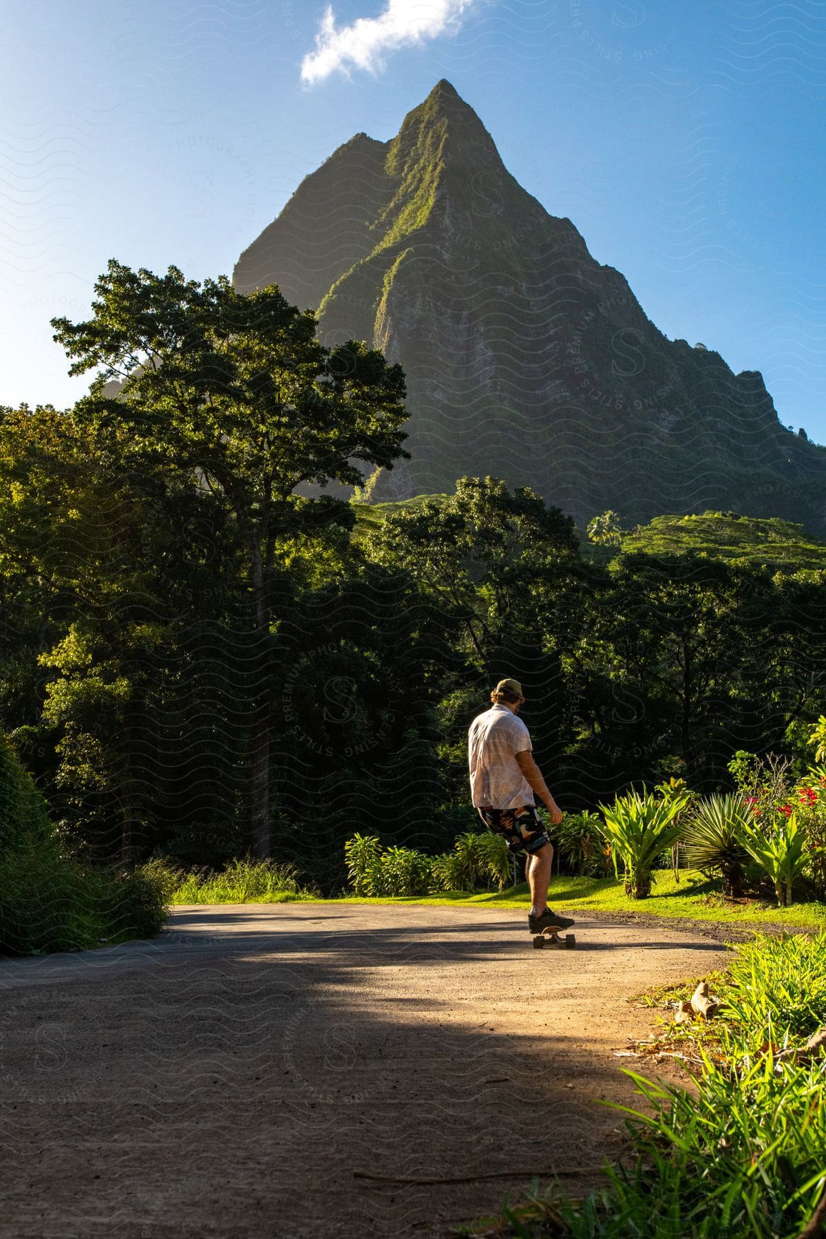 A Skateboarder Skates Along A Dirt Road Beneath A Green, Tropical Mountain