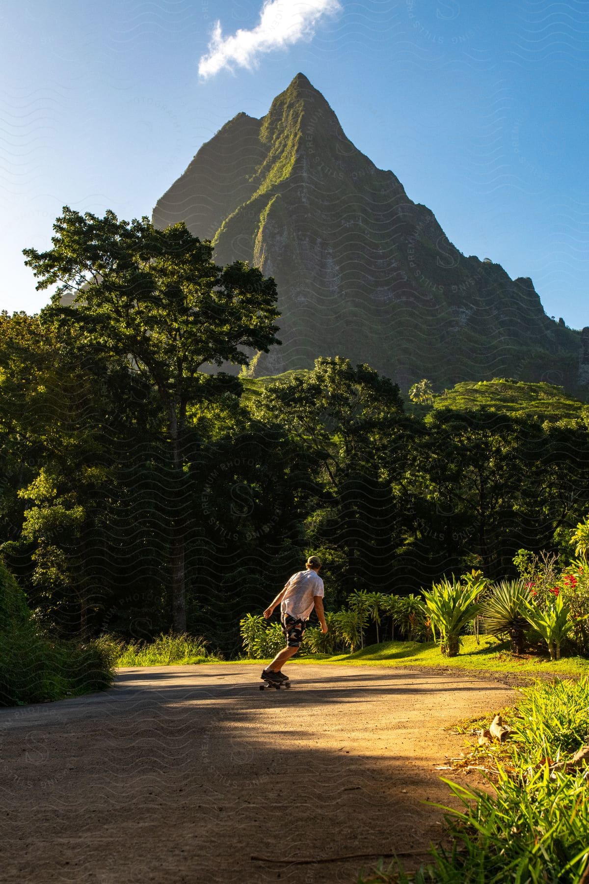 A man wearing a cap, a white T-shirt, and shorts skateboards along the dirt road with a mountain ridge in the background.