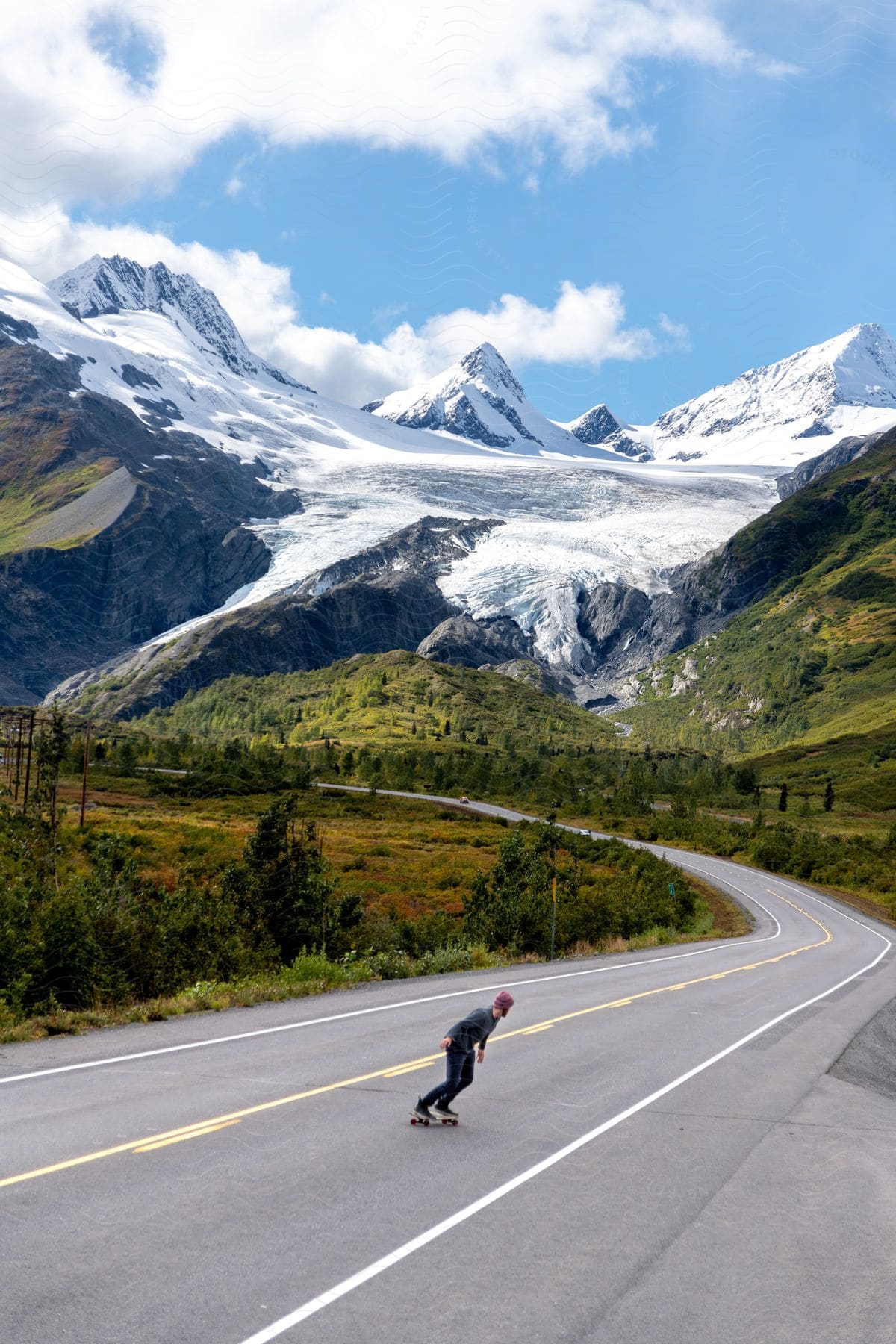 A person skateboarding on an asphalt street in a rural region with huge mountains covered in ice