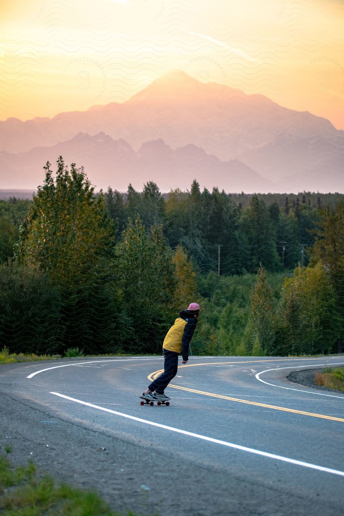 A person is wearing a black and yellow coat and pink helmet while riding a skateboard on an asphalt street and in the background there is a forest of trees