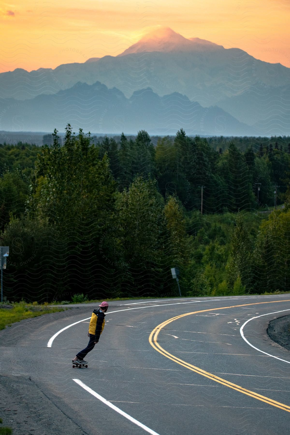 Man riding a skateboard on an asphalt near the vegetation of trees and mountain ranges on the horizon in the late afternoon sky.