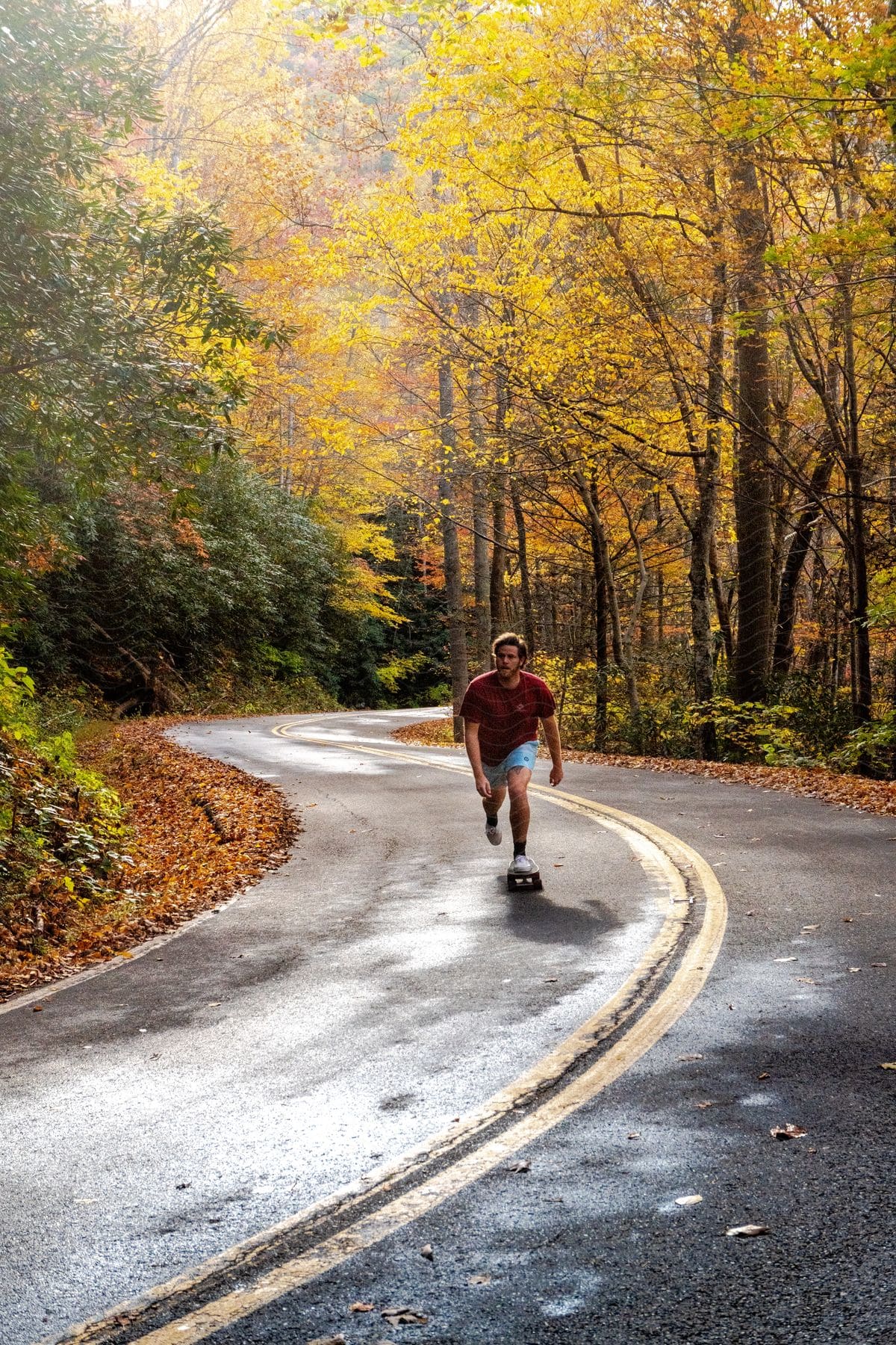 A man in a red shirt is boosting his skateboard on an asphalt street with lots of trees and vegetation on the sides