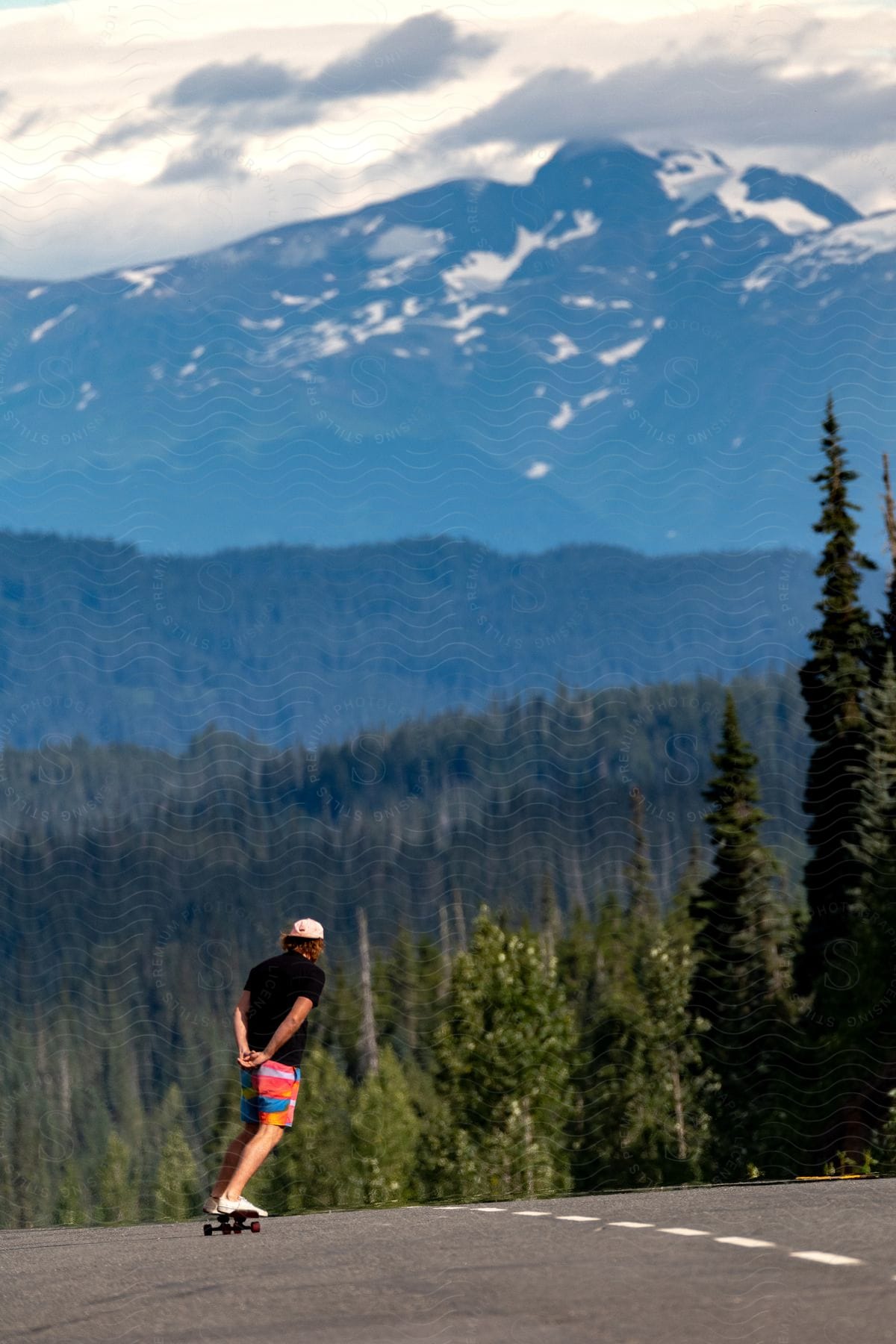 Man skateboarding on a mountain road with a forested landscape and snow-capped peaks in the background.