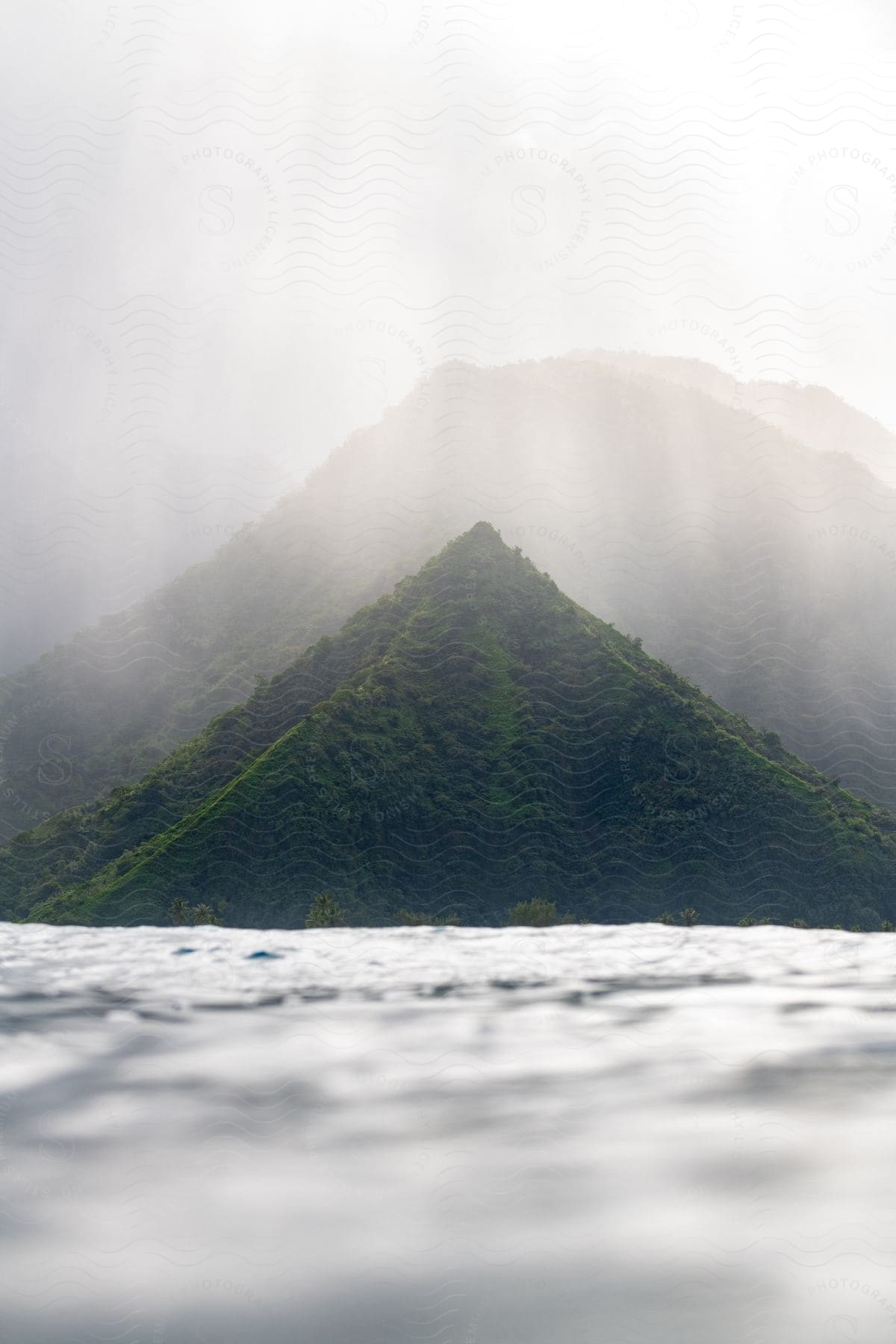 A large green mountain with vegetation in front of the sea and behind there is another mountain even bigger but it is hidden in the fog