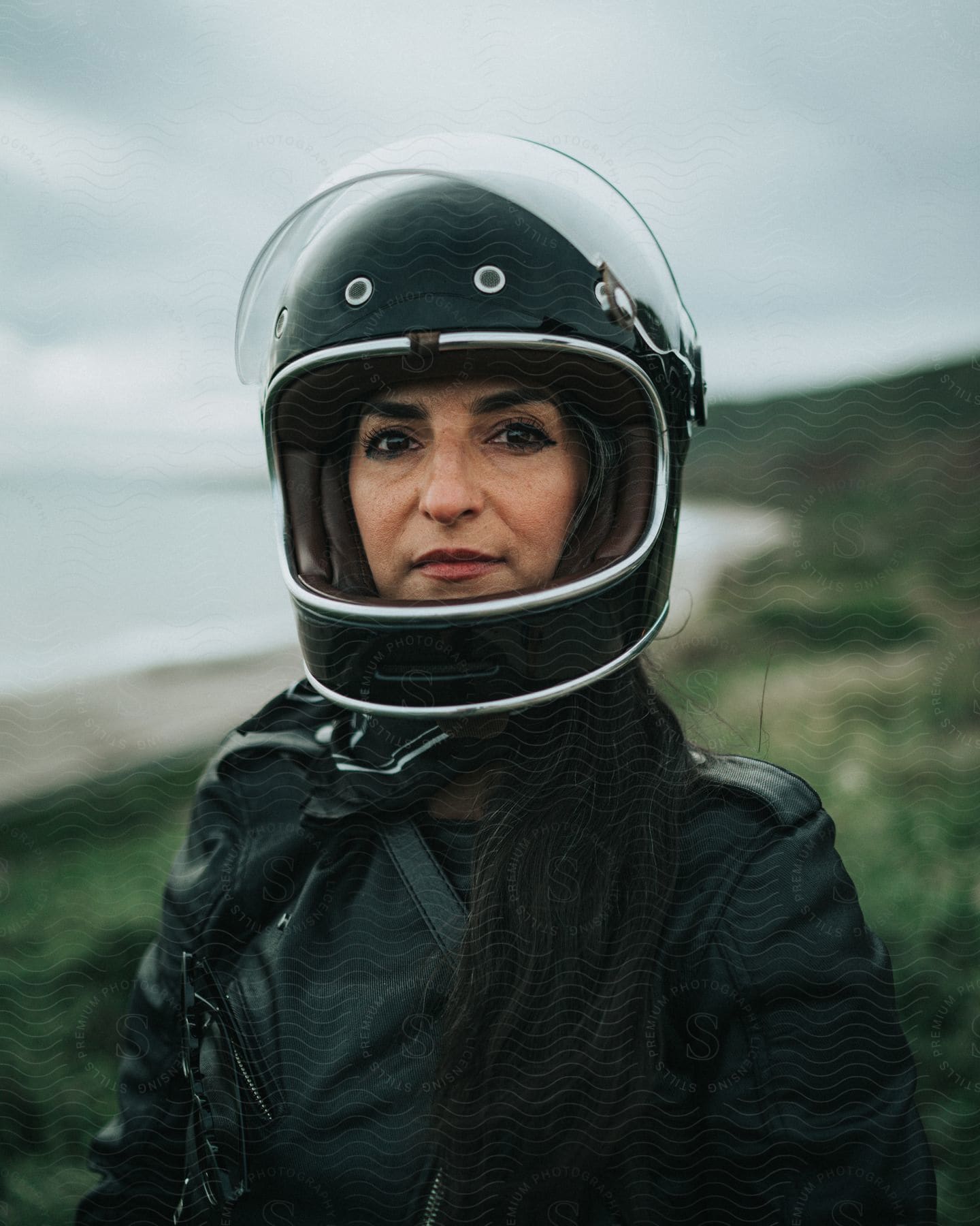 Woman wearing a black helmet and a black leather jacket stands on a hilltop with the coast in the background