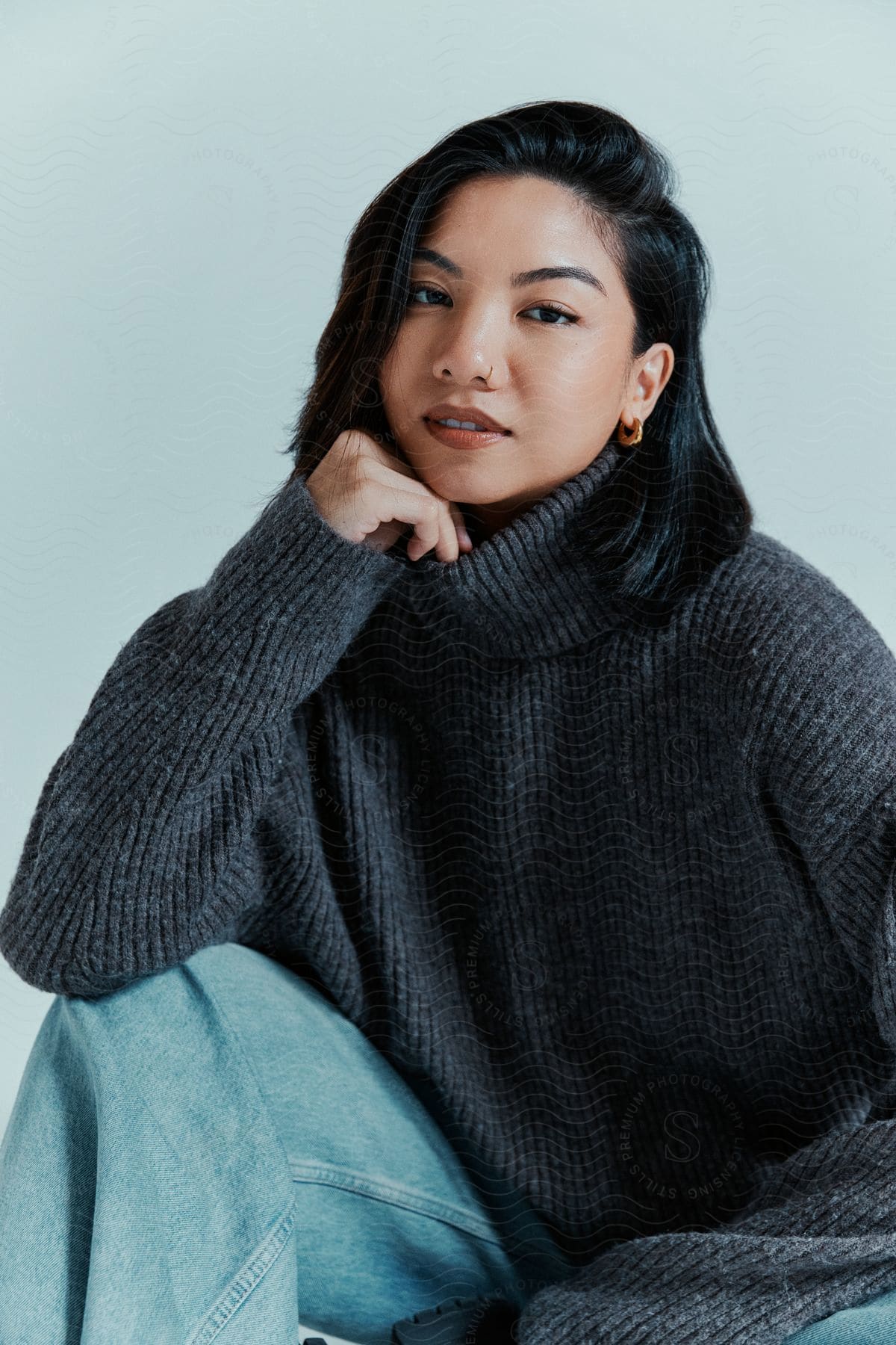 A woman is sitting wearing jeans and a gray sweater and her hand is holding her chin
