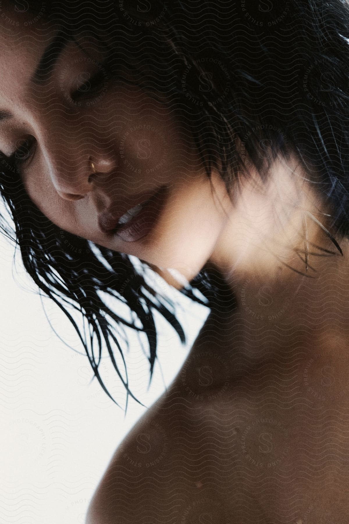 Close-up of a woman with wet, shoulder-length hair, a nose piercing, and light illuminating her chin and neck as she looks down.