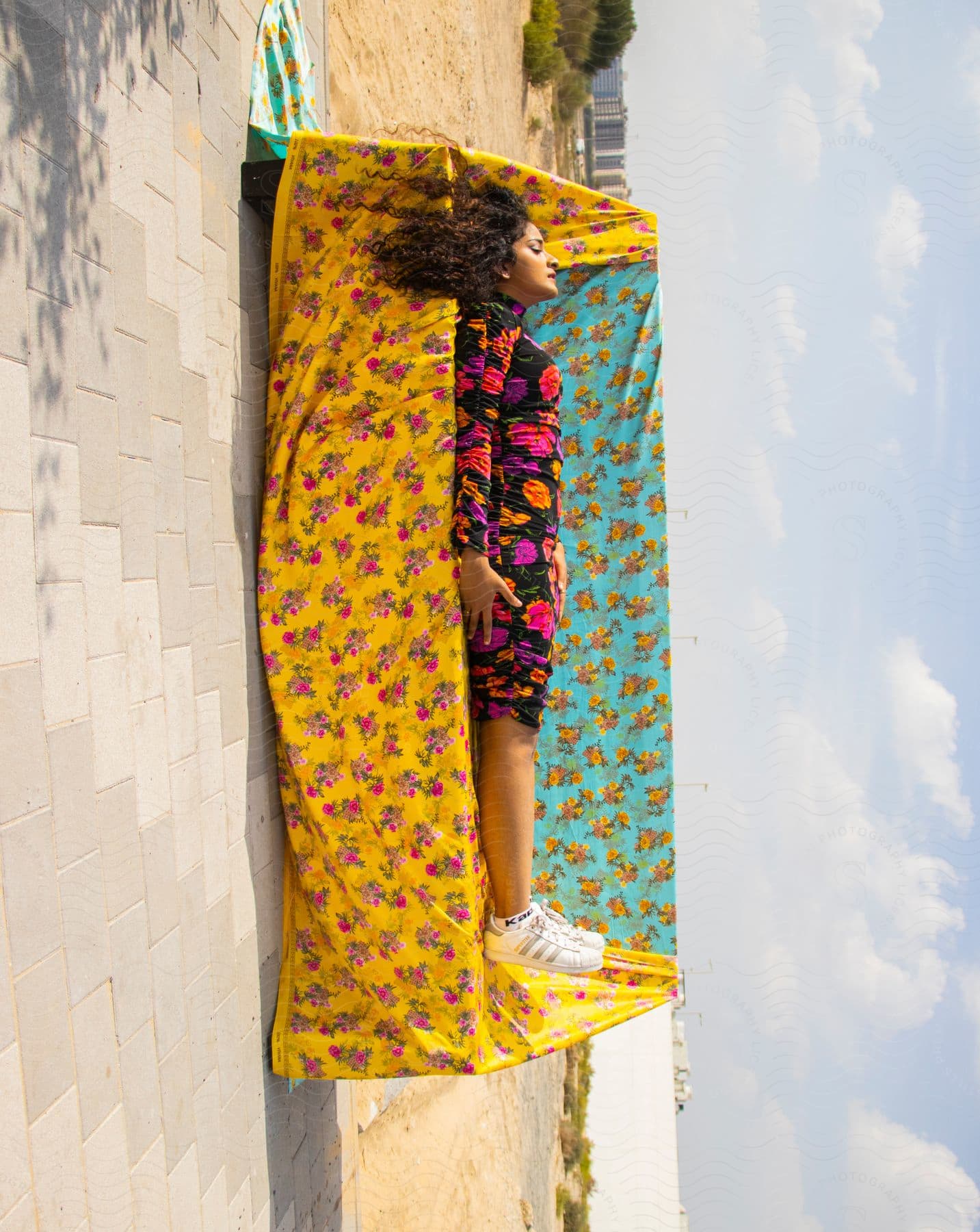 Woman relaxes lying on a bench patterned with yellow and blue floral fabrics outdoors on a blue sky day.