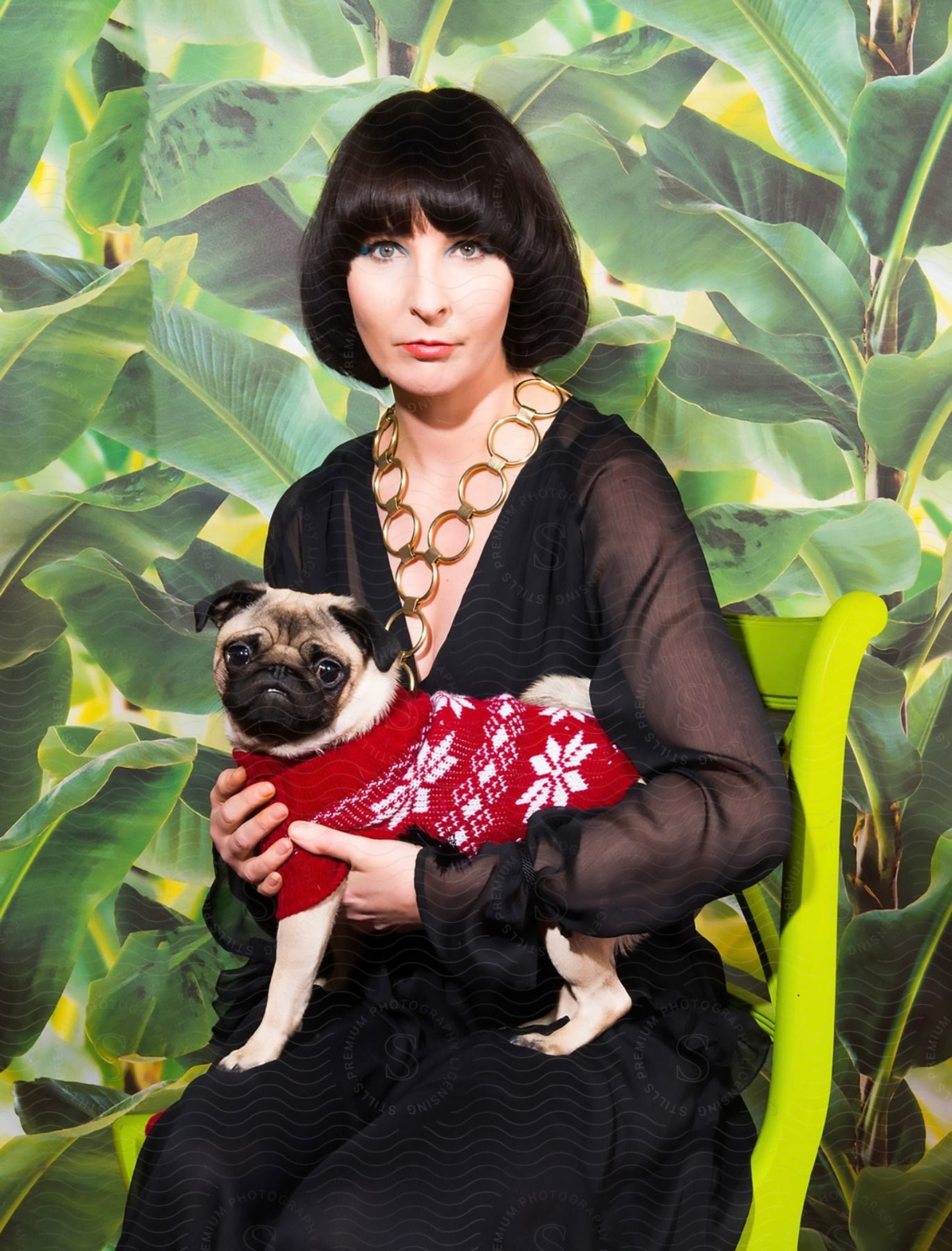 A woman holds a dog wearing a red sweater, with large green leaves in the background.