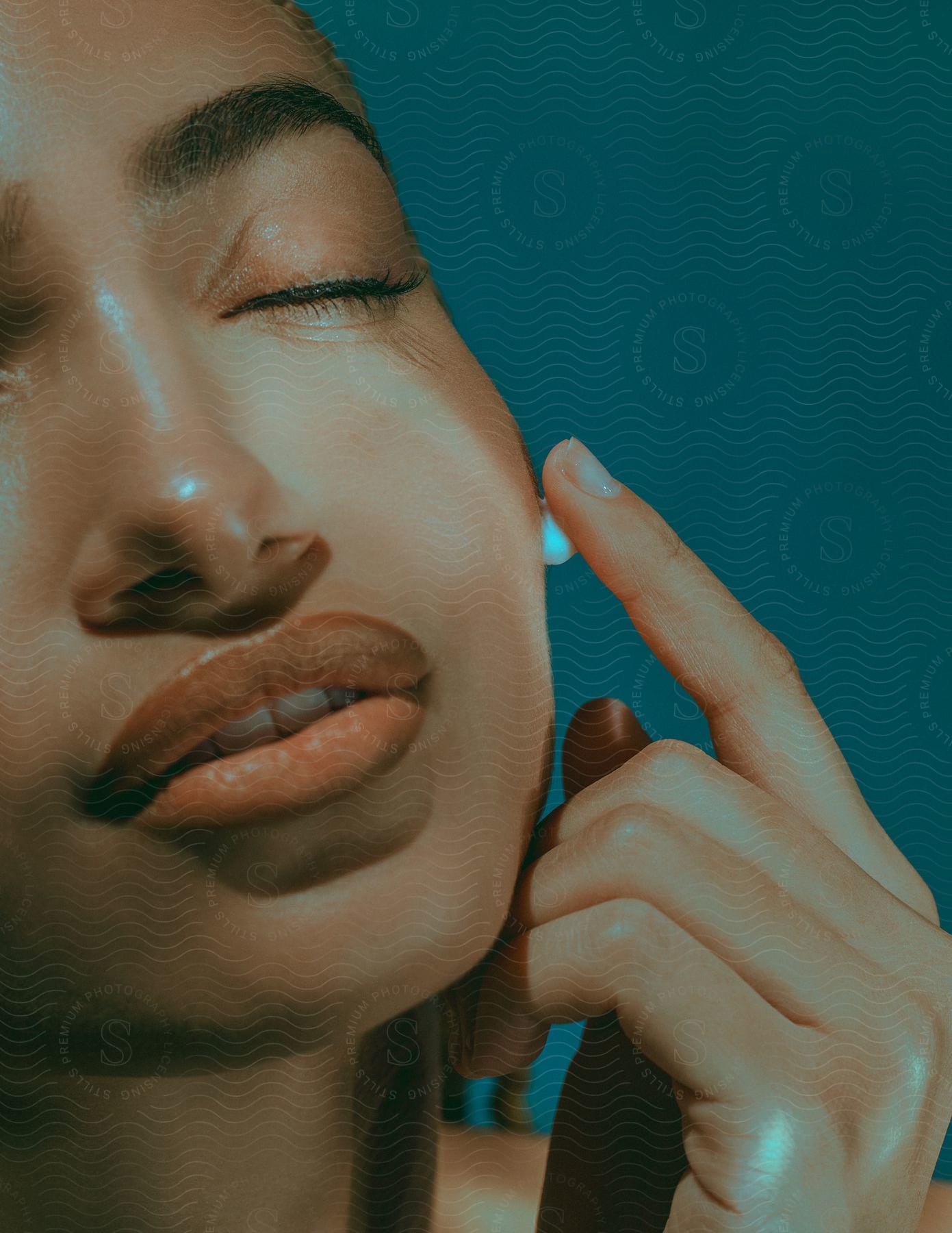 Close-up of a woman's face with her eyes closed and finger placed on her cheek applying a product, against a blue background.