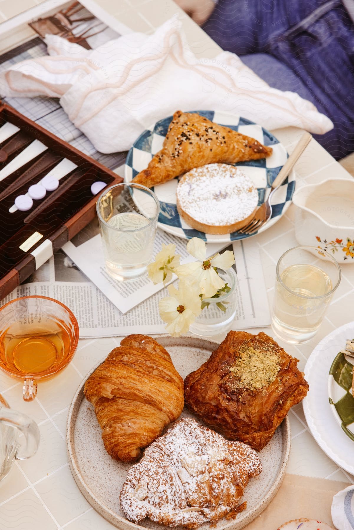 A table with a variety of pastries and drinks, accompanied by a backgammon board. Blooming white flowers decorate the setting, creating a relaxed atmosphere suitable for a casual meal or date.