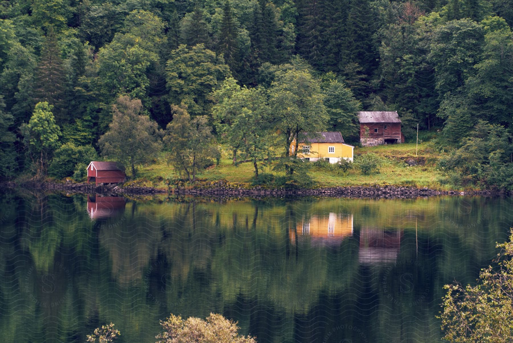 A serene landscape with a tranquil lake surrounded by green forests and a few buildings at the water's edge. The reflection of the trees and buildings in the calm lake water creates a mirror effect.