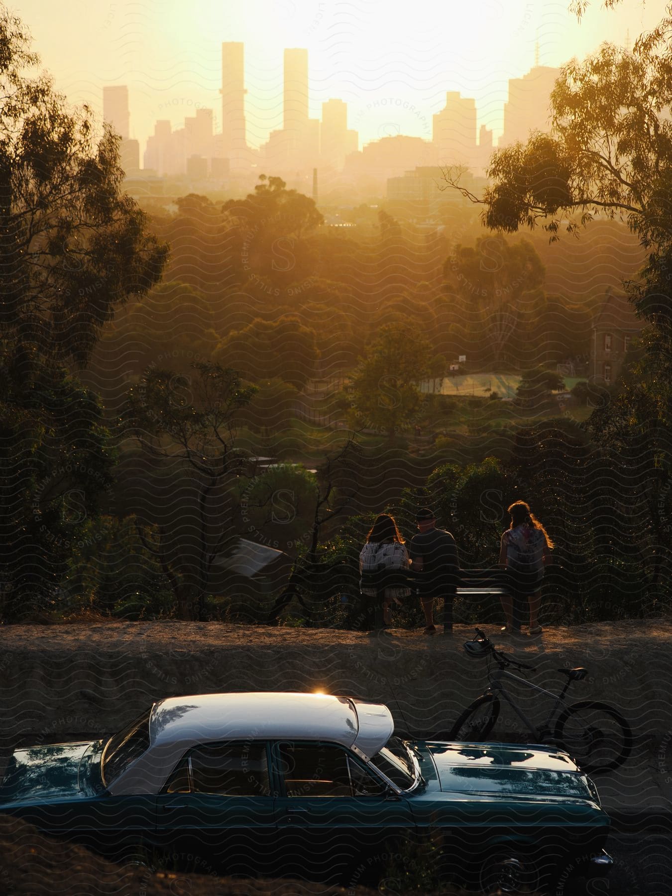 Three people sitting on a bench and behind there is a bicycle and a car parked on the horizon there are city buildings illuminated by a sunset