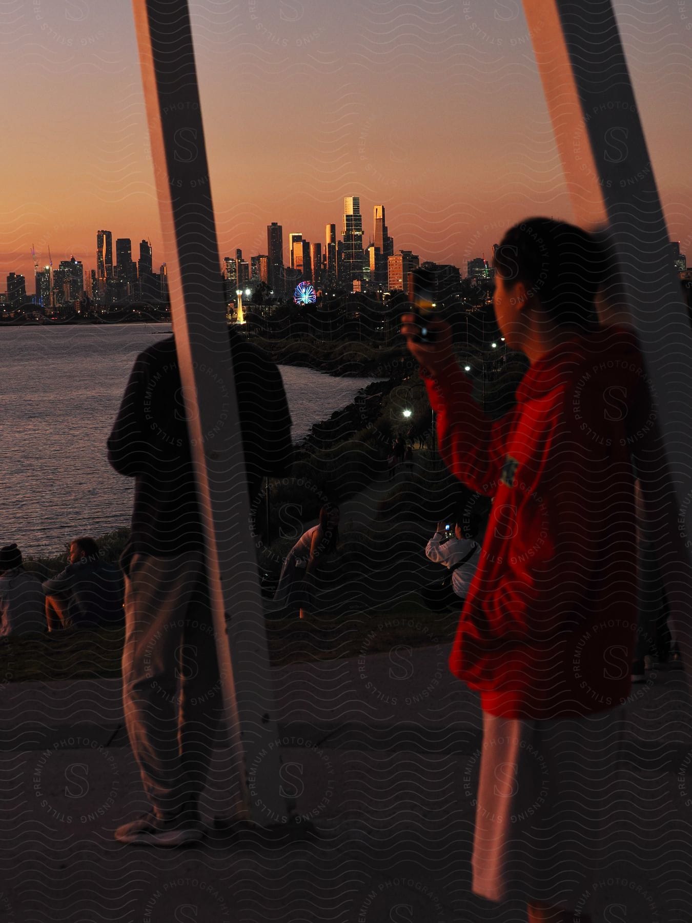 During sunset, people enjoy the view of a city skyline from a park.