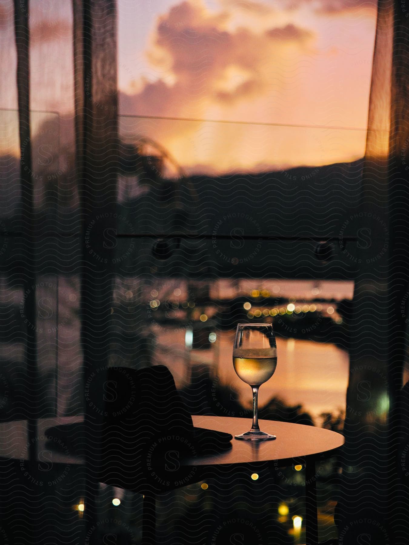 Glass Of Wine On A Table With A Fedora Hat Against A Backdrop Of A Sunset Viewed Through Sheer Curtains