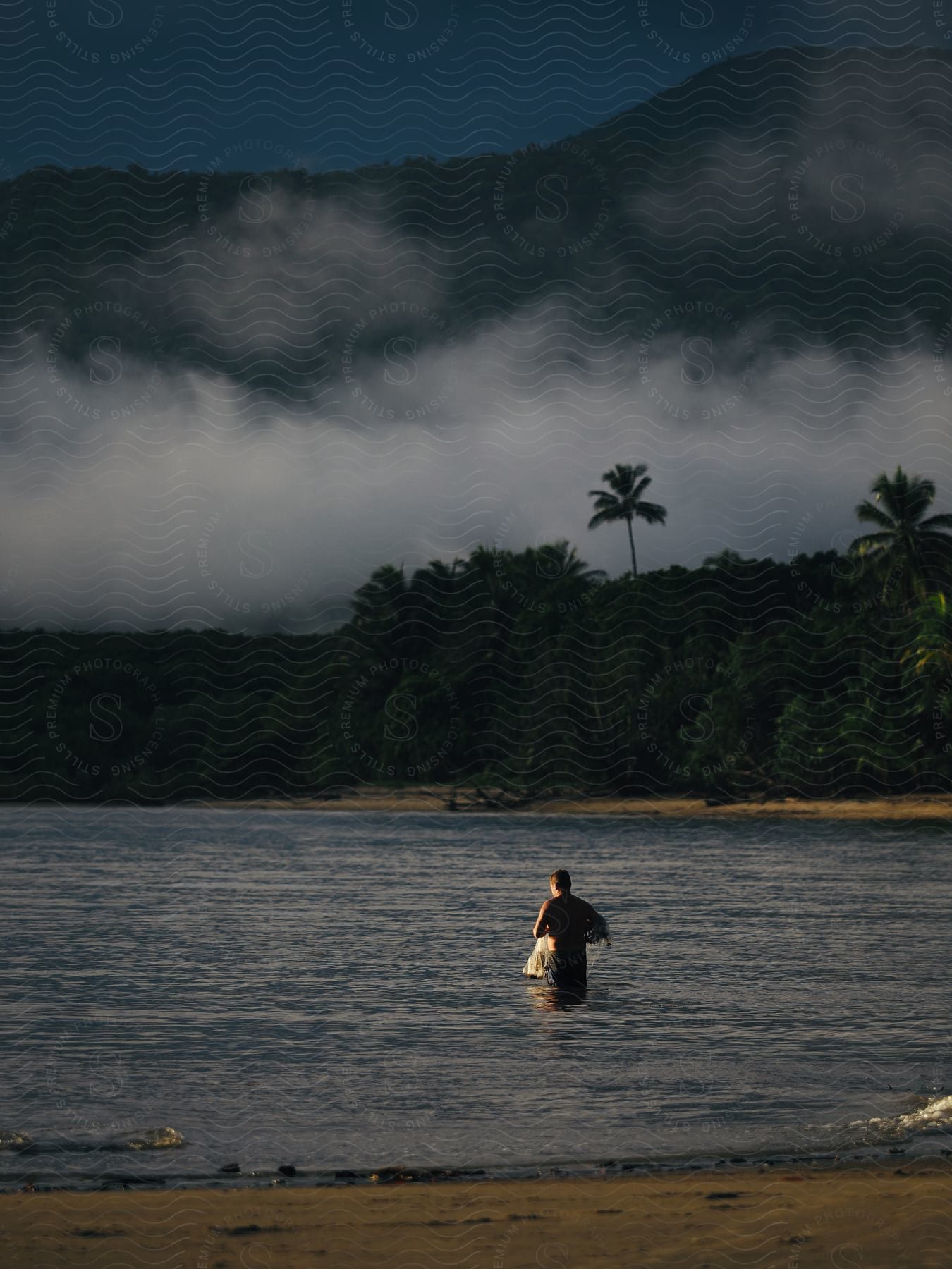 A man stands in shallow ocean water at sunset on a cloudy day.