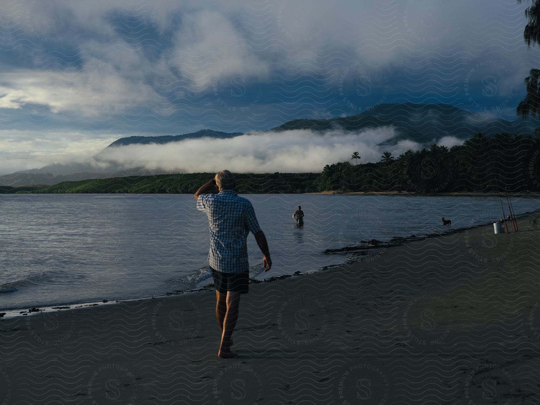 Man walks on a beach as another person and a dog are in the water with mountains in the distance