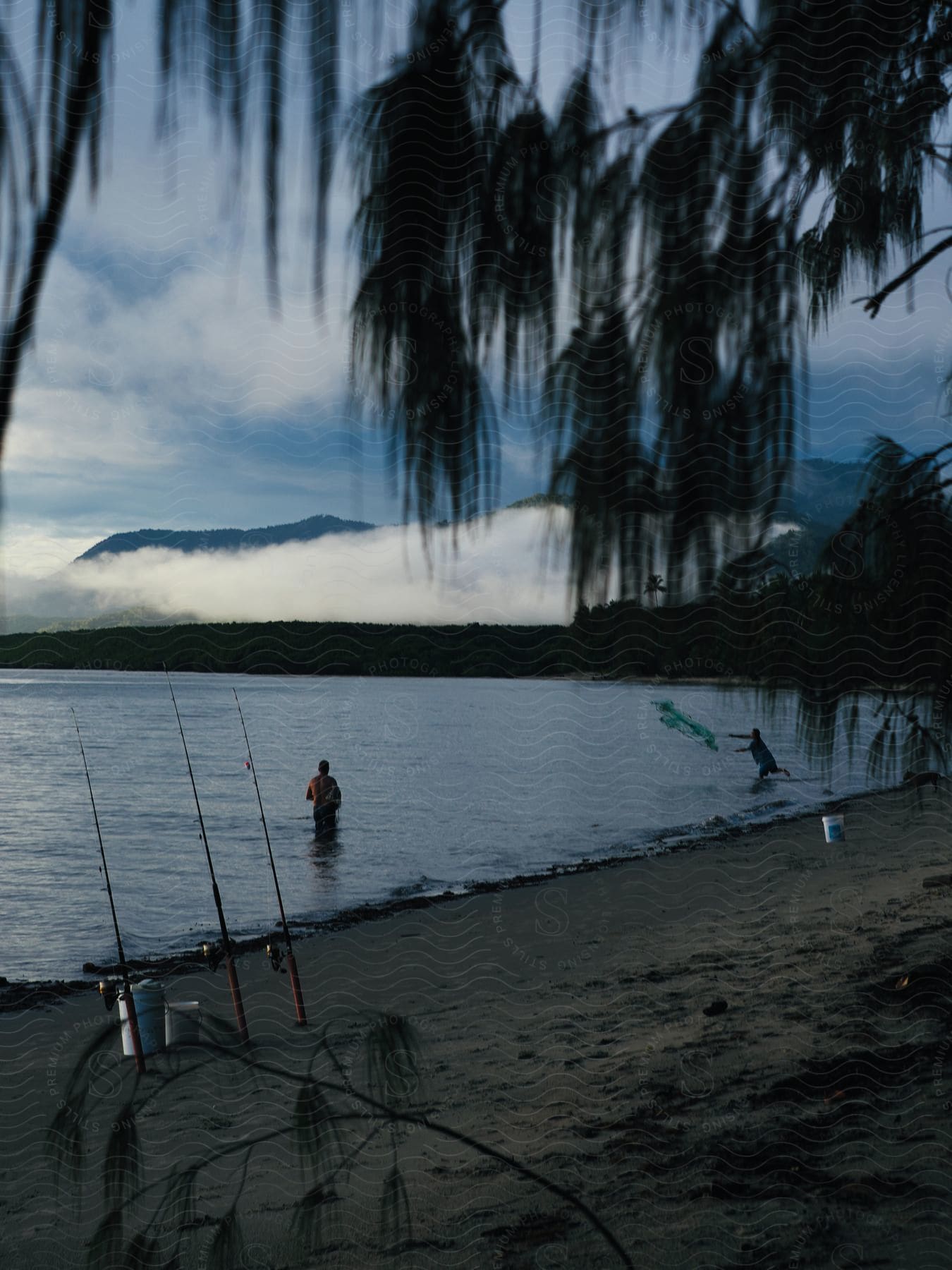 Landscape of a beach with mountains around and fishing rods with two fishermen by the sea on a cloudy morning.