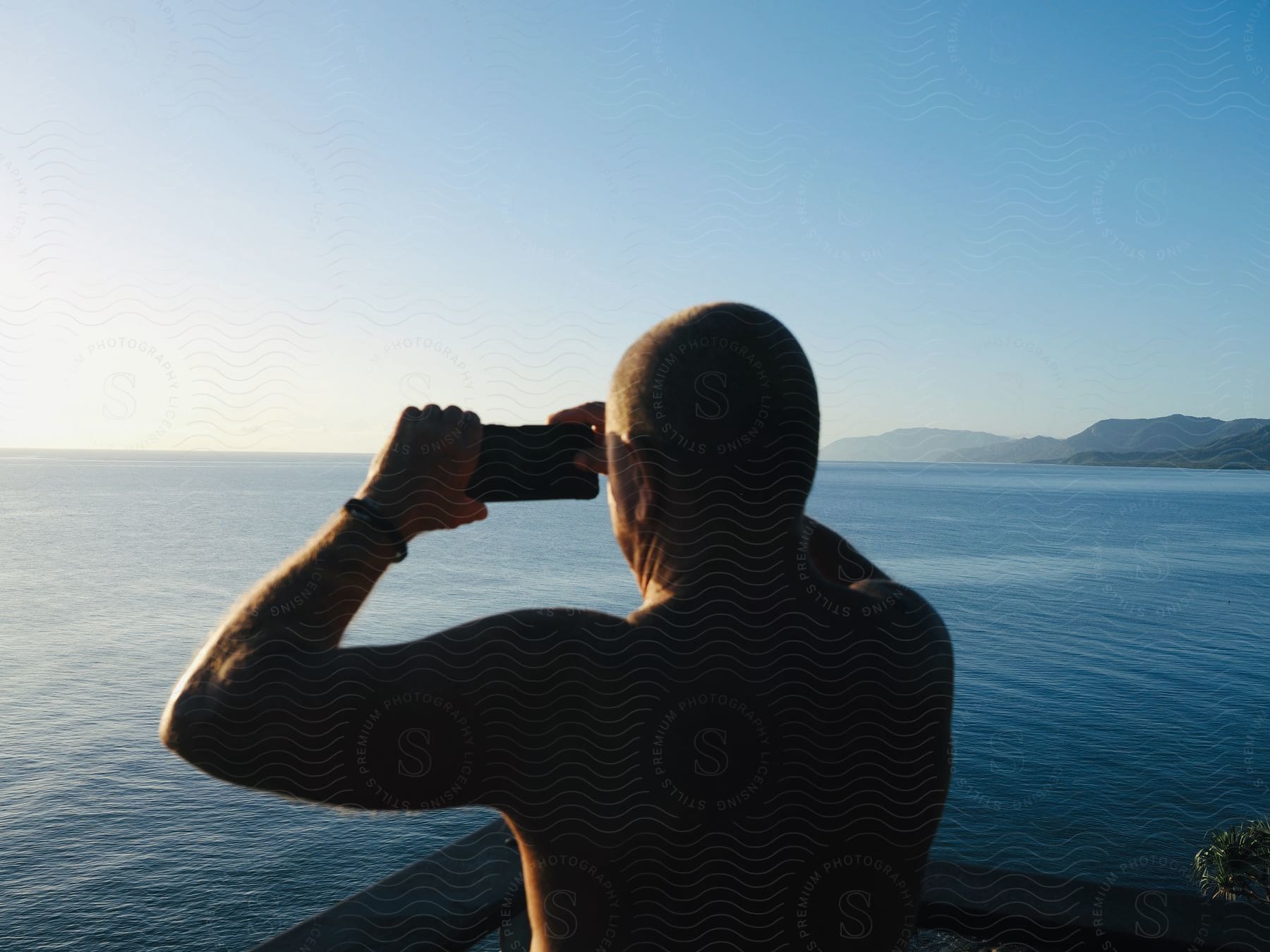 A shirtless man standing on a deck above the water taking a photo with his cell phone camera