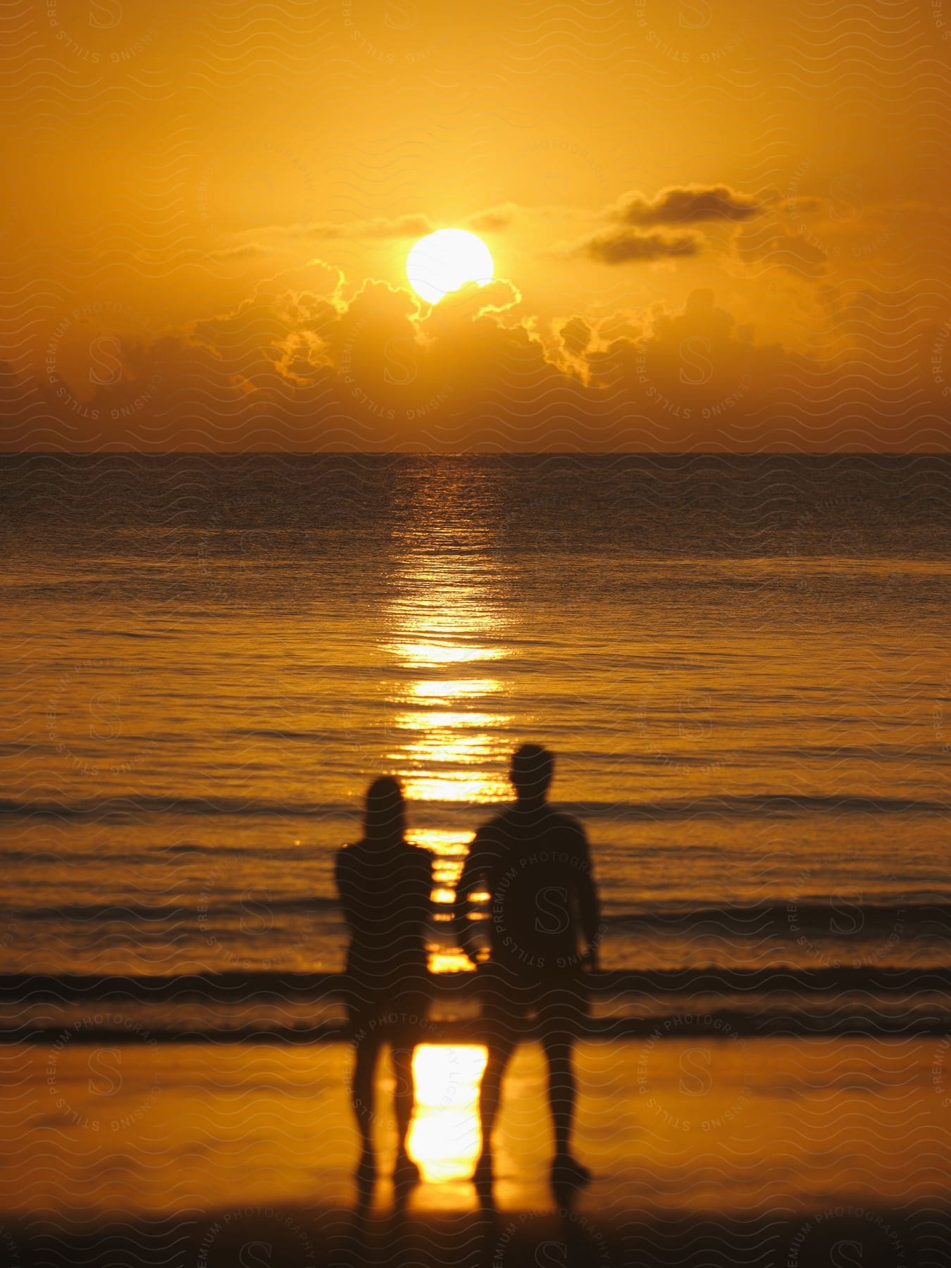 A silhouetted couple stands atop a beach, basking in the glow of a sunset descending over the ocean.