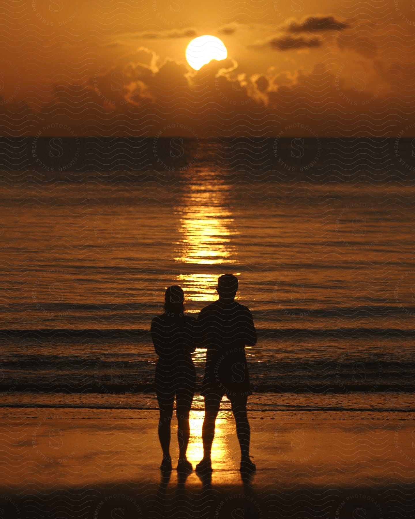 A man and woman stand on the ocean shore at sunset.