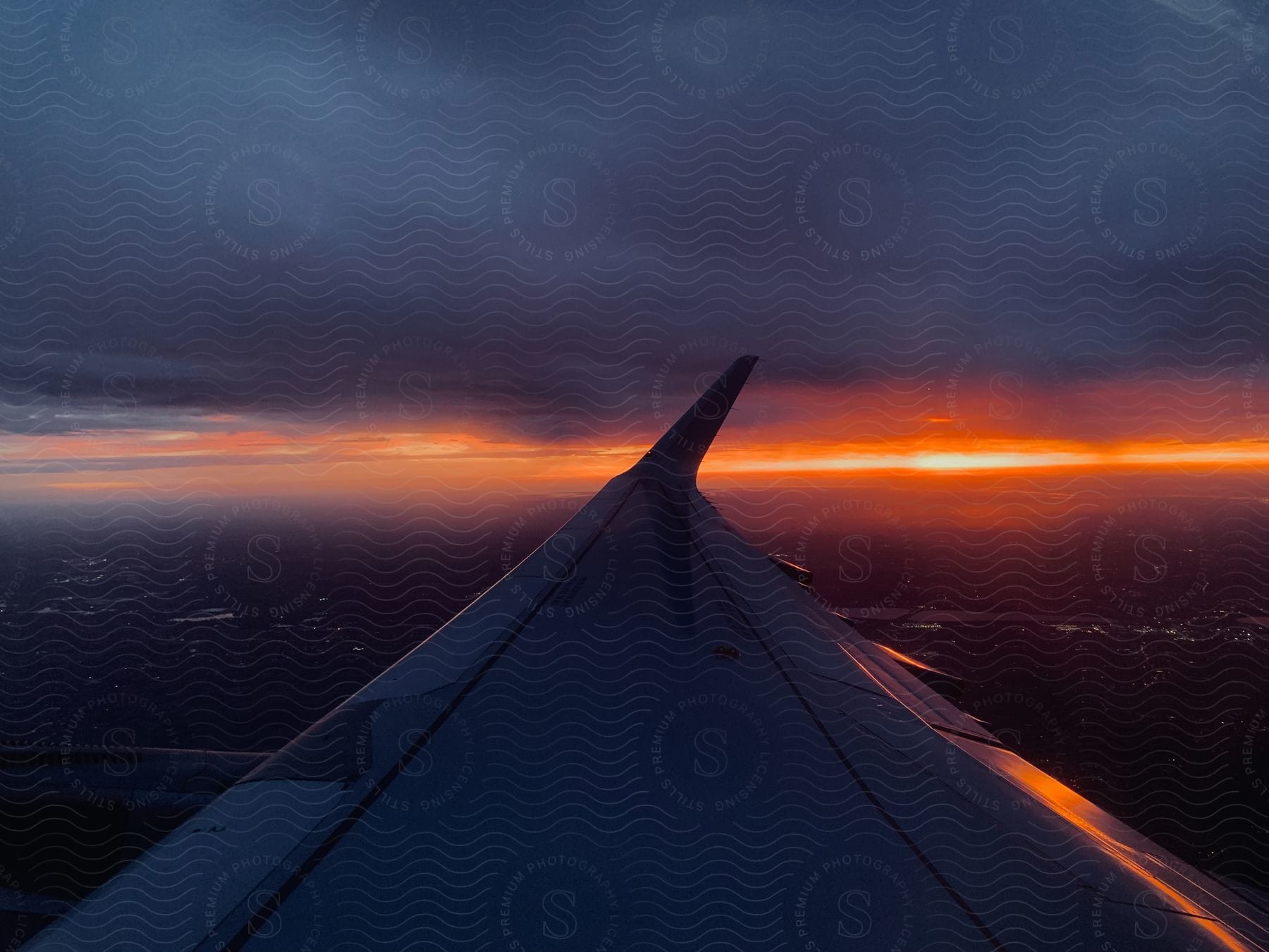 The wing of a passenger jet flying through the sky at dusk with a strip of bright orange sun under gray clouds