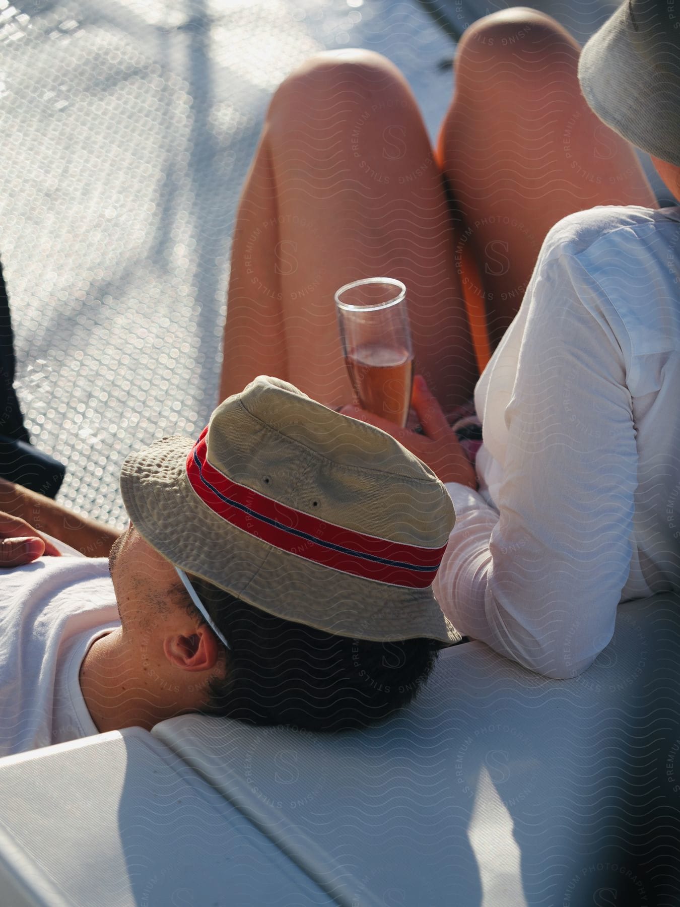 A man in a hat relaxes on a white tarp mat on a sailboat, while a woman nearby holds a champagne flute and wears a hat and a sleeveless shirt.