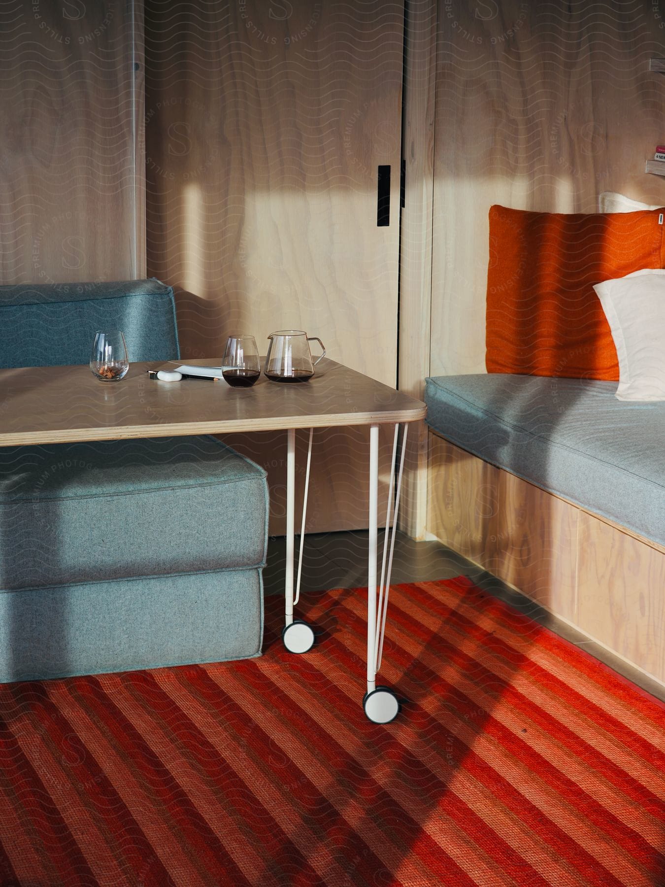 A cozy room furnished with a bed, a couch, and a white-wheeled table holding a drink and a classic pitcher. A red and orange striped rug warms the floor.