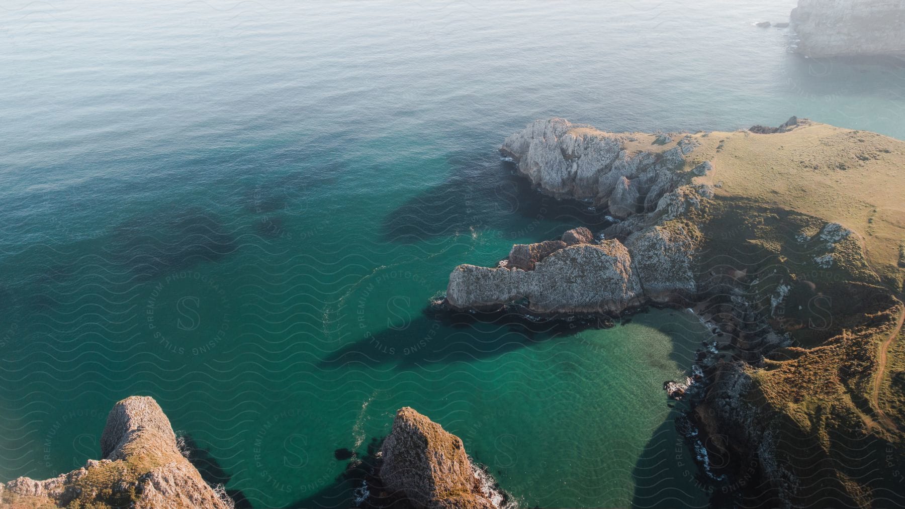 An aerial view of a rocky coastline, surrounded by blue-green waters. The rock formations emerge irregularly from the water, while the land is covered in green grass.