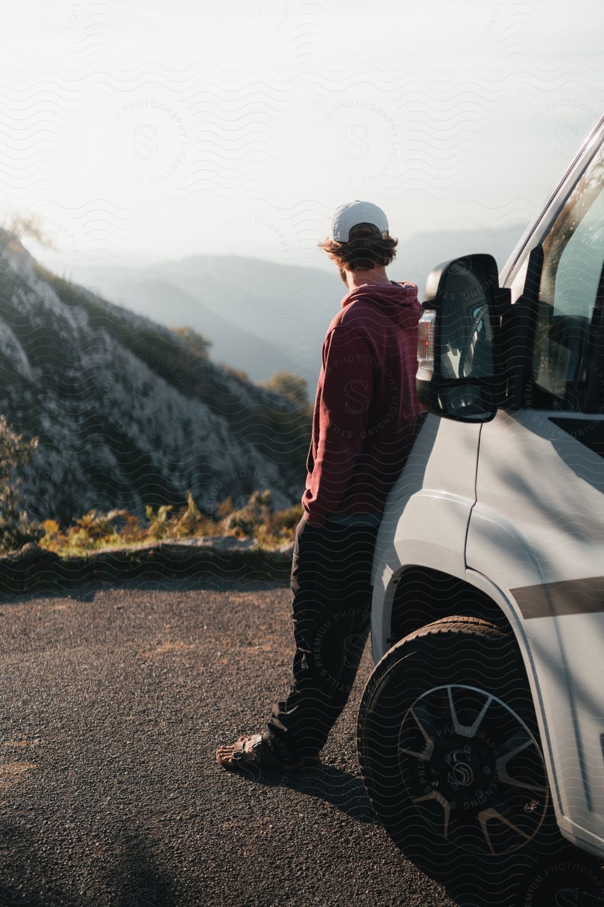 A man wearing a white cap, a red hoodie, and dark pants leans against a van, watching the mountainous scene.