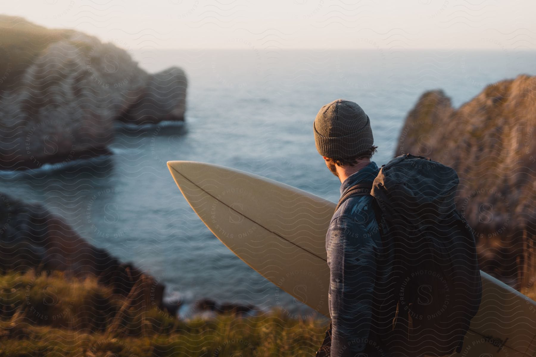 Man holding his surfboard and with a backpack on his back walking on the coast near the sea.