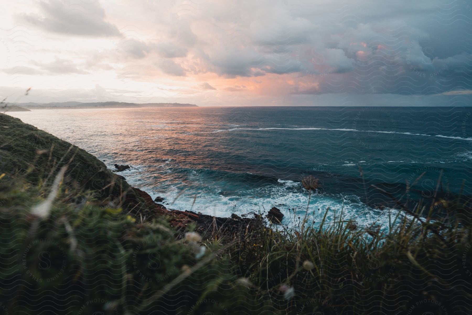 A coast of a sea with small waves crashing on the rocky shore and There are many clouds in the sky with an orange color from the sunset