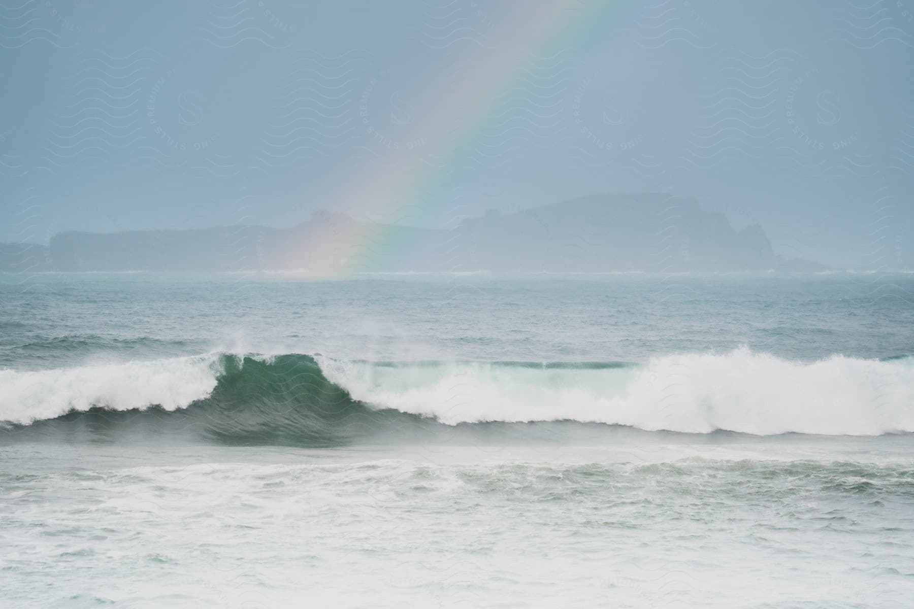 A sea wave breaking on the surface of the water and behind it is a rainbow