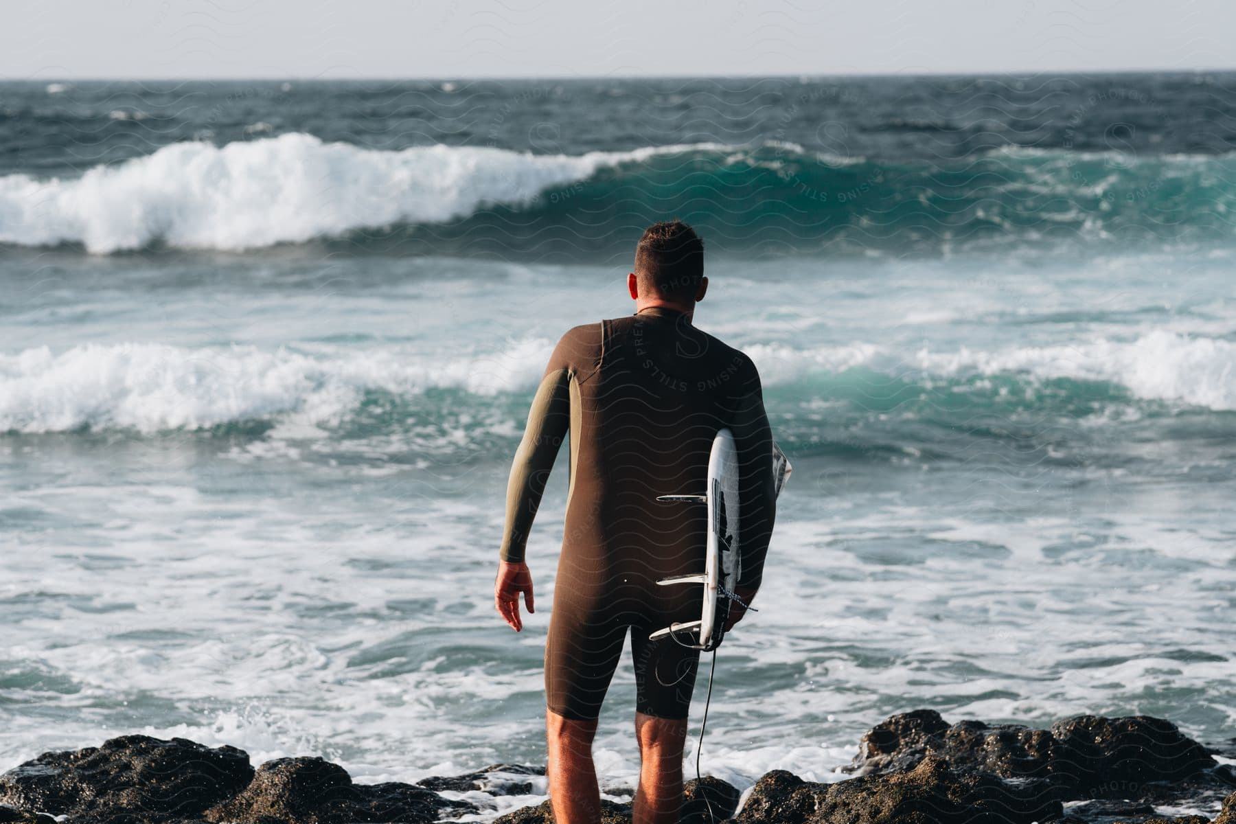 Man with his back turned and surfer standing on the rocks, watching the sea waves while holding his surfboard.