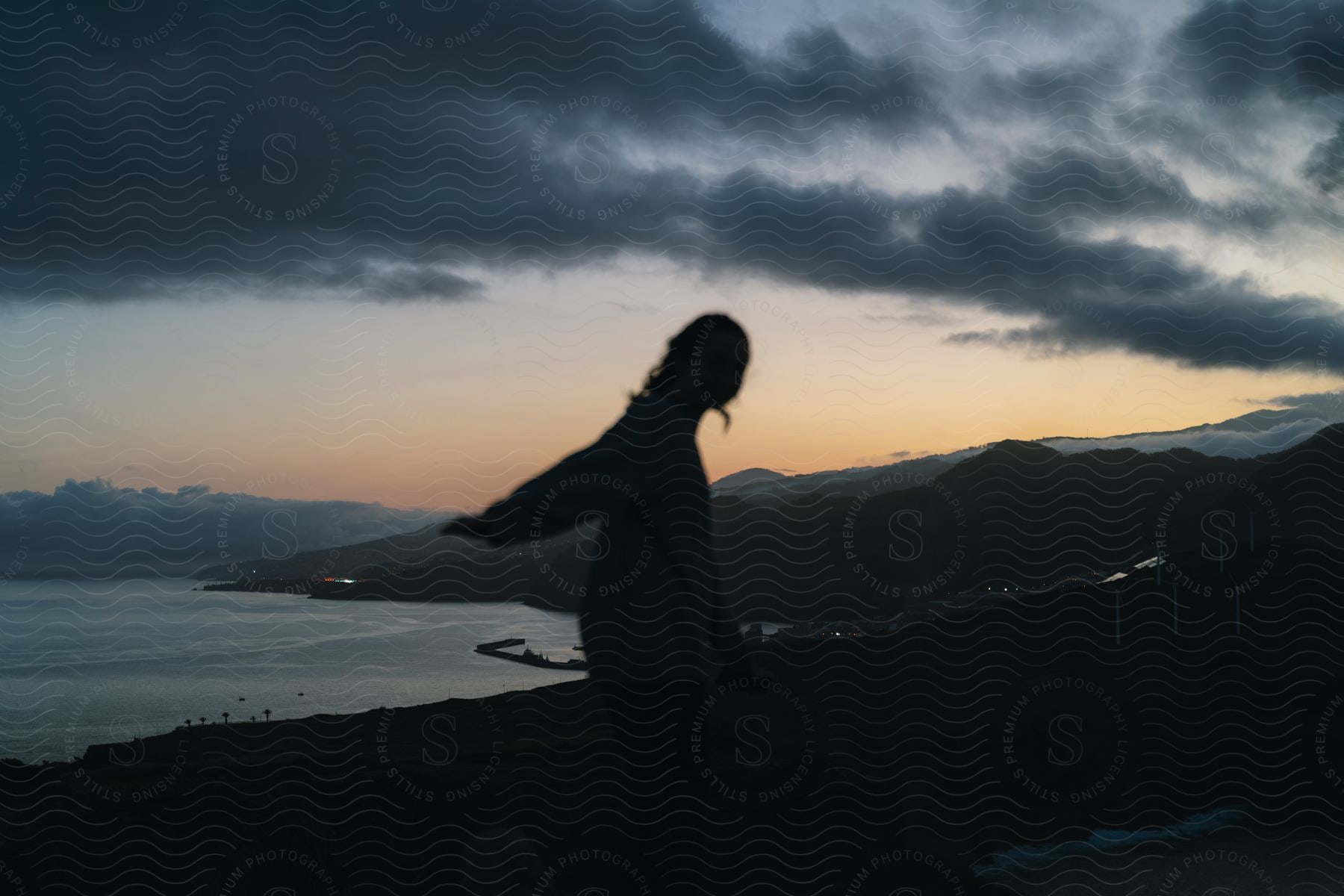 Silhouette of a person standing along the coast with mountains in the distance under a dark cloudy sky