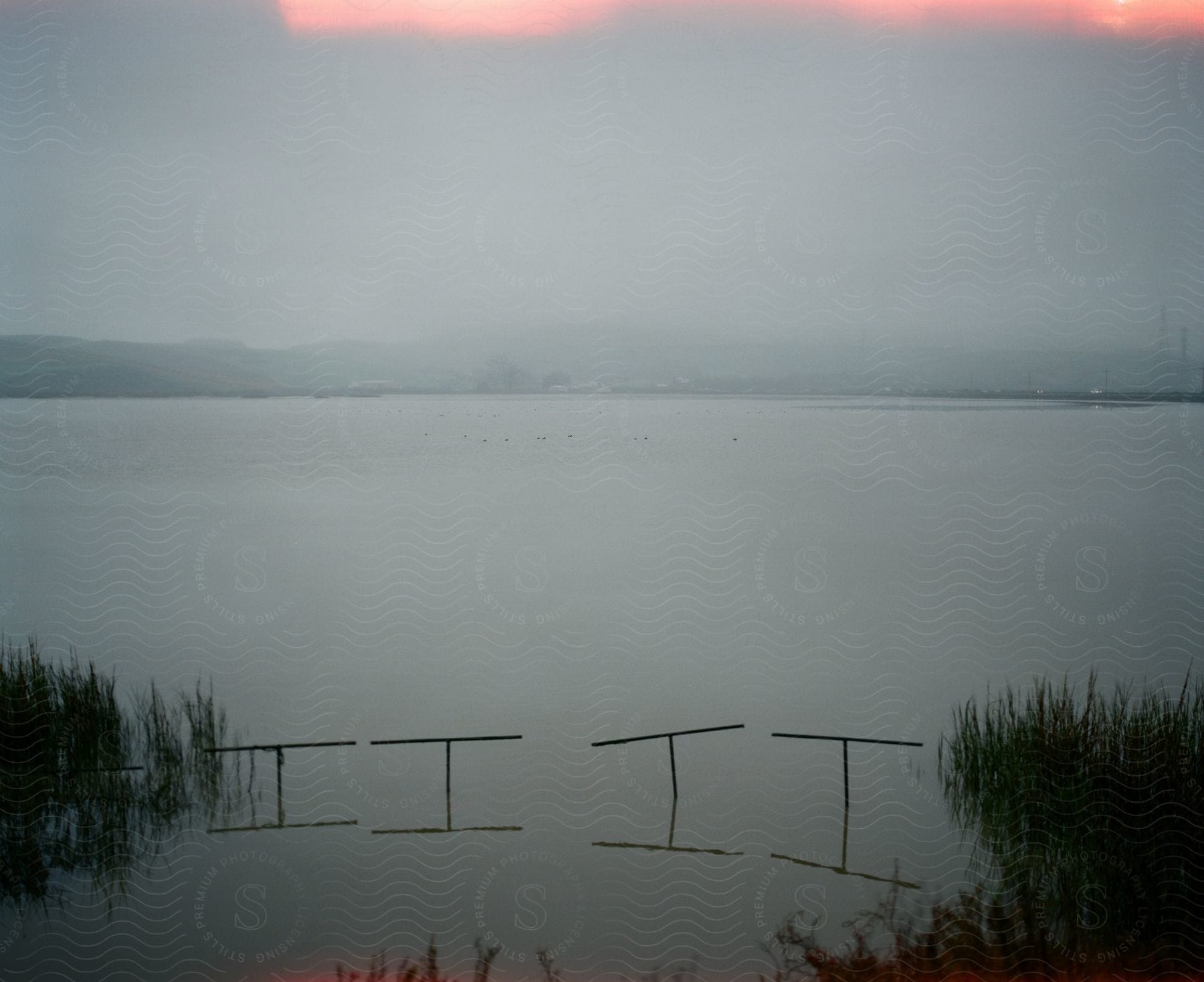 Posts with perpendicular top pieces sit near the shore across the water from a fog covered town on an overcast day.