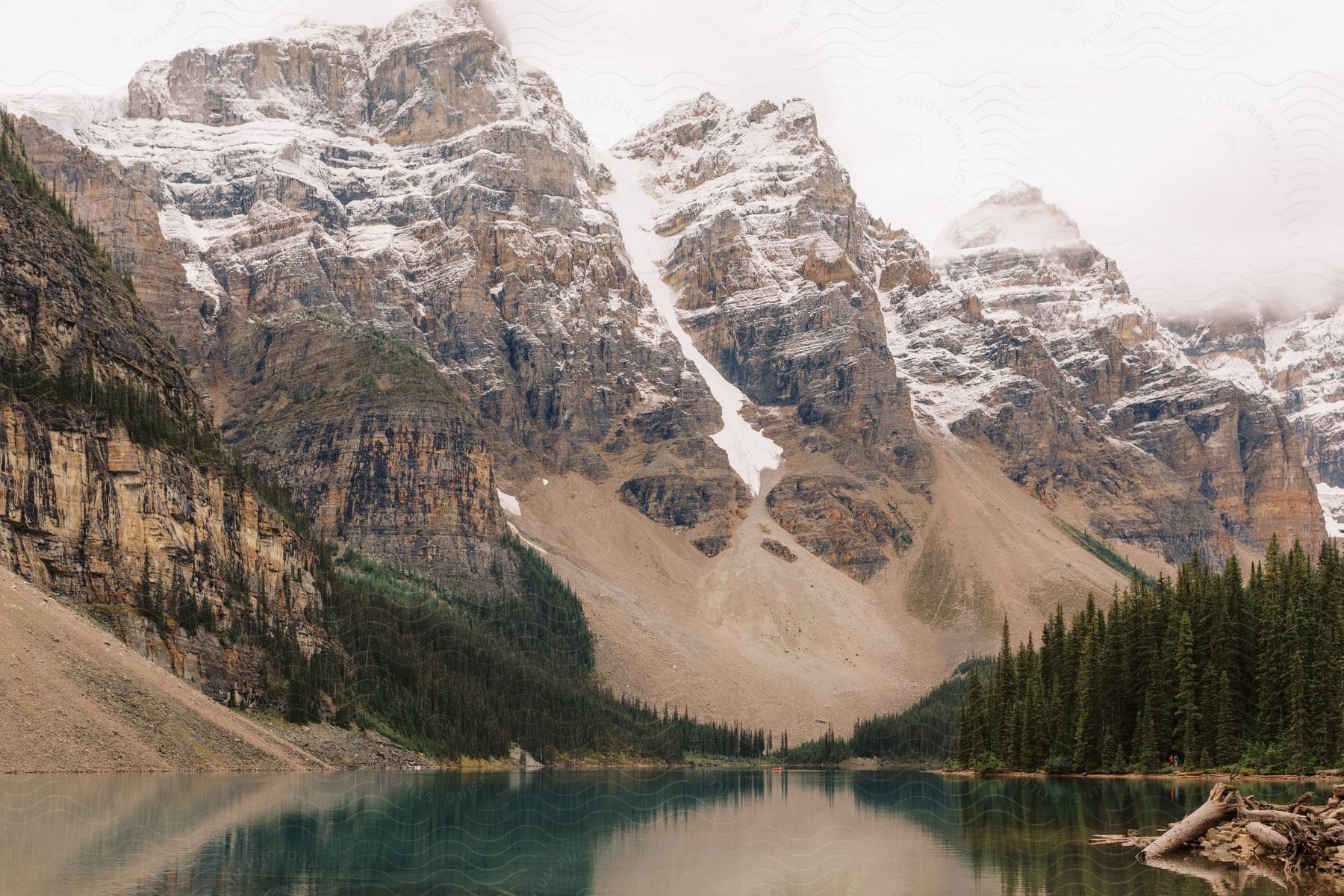 Lake reflects snowy mountains and conifer forest on a cloudy day.