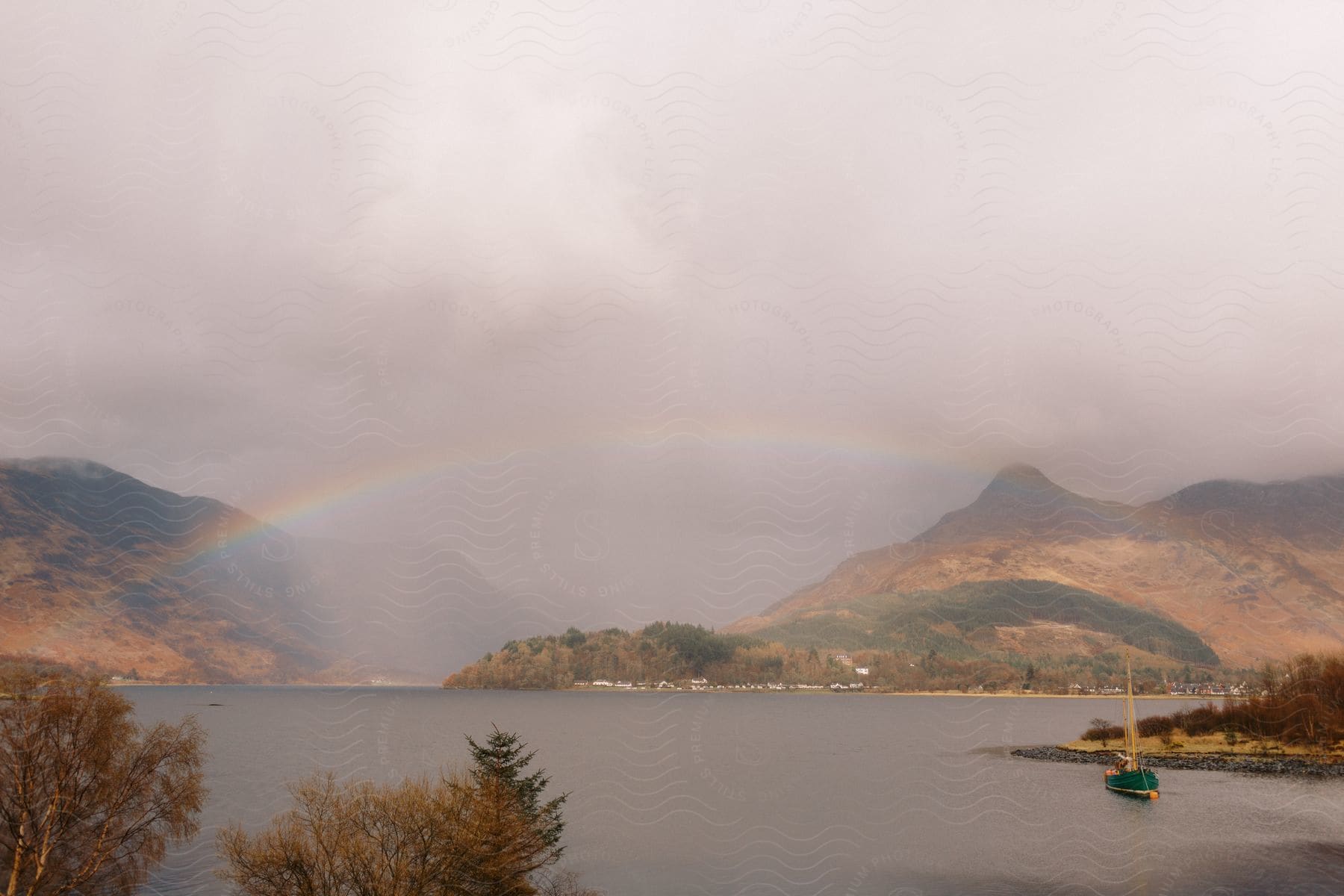 A boat floats along the river near the riverbank on a cloudy day, with a rainbow arching over a mountain in the background.