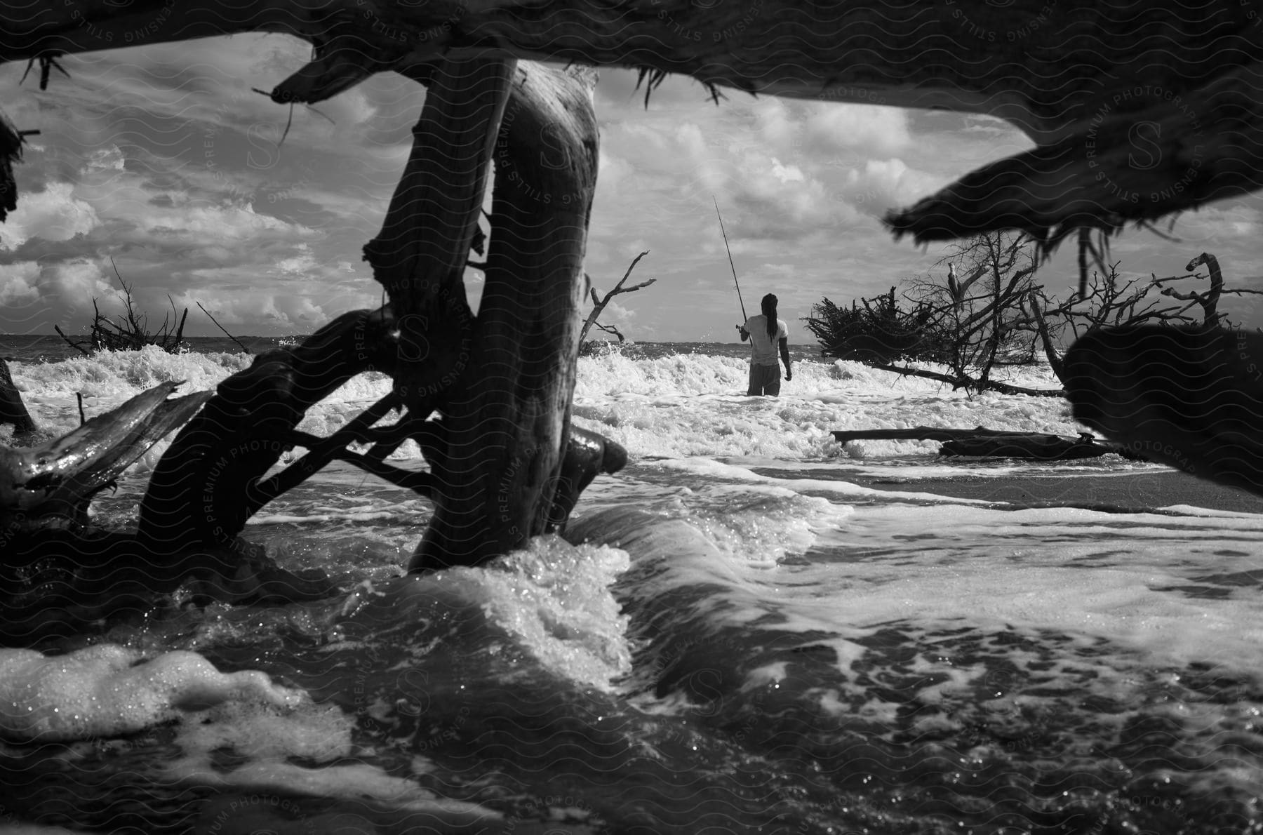 A man fishing on the coast amidst piles of driftwood.